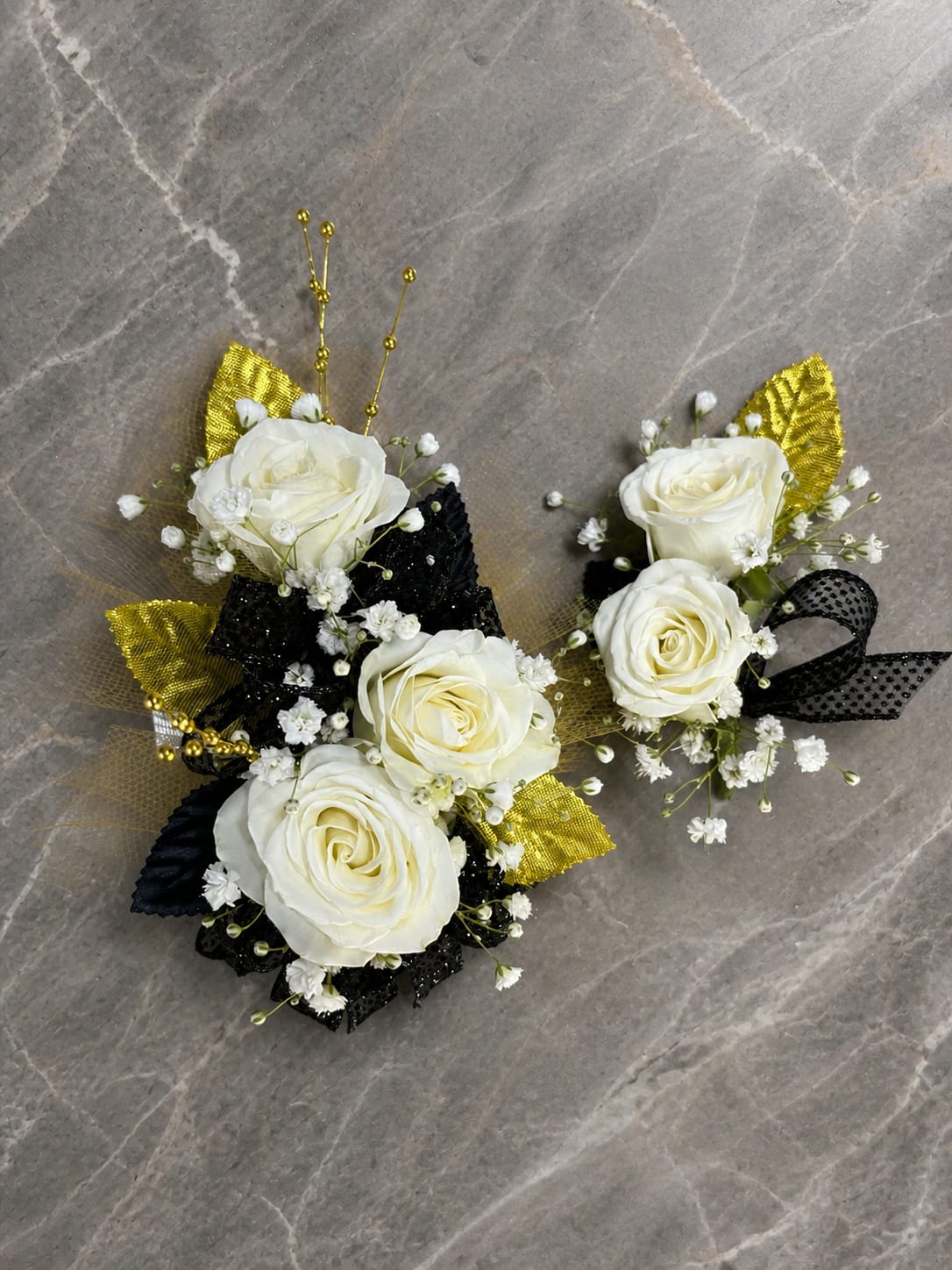 Formal Affair Set - MAGNETIC BOUTONNIERE WARNING: CONTAINS STRONG MAGNETS. DO NOT USE IF YOU HAVE A PACEMAKER, DEFIBRILLATOR, INSULIN PUMP, COCHLEAR IMPLANT, NEUROSTIMULATOR, OR OTHER IMPLANTED MEDICAL DEVICE. KEEP AWAY FROM ELECTRONICS, CREDIT CARDS, HOTEL KEY CARDS, AND CHILDREN.  Formal Affair  White roses, black ribbon, gold accents, and delicate filler flowers come together in Formal Affair, a sophisticated corsage and boutonniere collection designed for prom, homecoming, dances, banquets, weddings, and formal events. The timeless combination of black, gold, and white creates a polished black-tie look that is both elegant and unforgettable.  Standard includes a wrist corsage with 3 spray roses and a matching magnetic boutonniere with 2 roses. Deluxe includes 4 spray roses in the corsage. Premium includes 5 spray roses for additional fullness and flower coverage. All boutonnieres include 2 roses.  Perfect for prom flowers, wrist corsages, magnetic boutonnieres, corsage and boutonniere sets, homecoming flowers, dance flowers, and formal event flowers.  Because each corsage and boutonniere is individually designed, no two sets will be exactly alike. Ribbon, filler flowers, bracelets, decorative accents, and other design elements may vary based on availability. While individual details may differ, the overall color palette, roses and/or flowers, style, and appearance of the Formal Affair collection will be maintained.