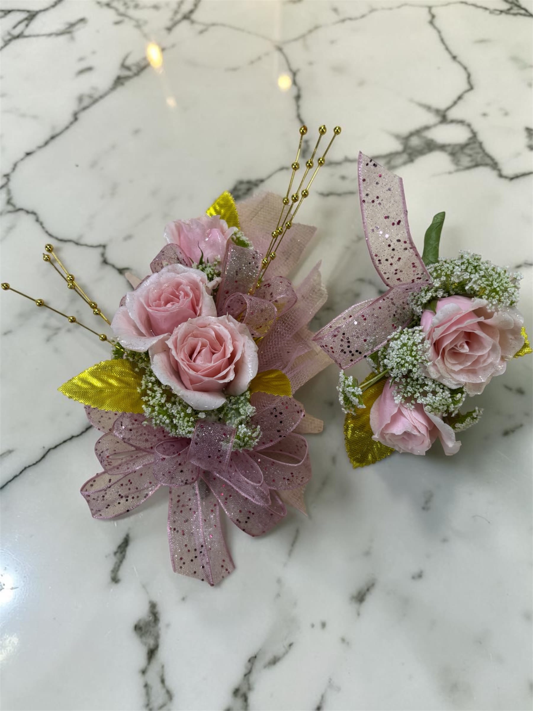 Rise Gold Formal Set - MAGNETIC BOUTONNIERE WARNING: CONTAINS STRONG MAGNETS. DO NOT USE IF YOU HAVE A PACEMAKER, DEFIBRILLATOR, INSULIN PUMP, COCHLEAR IMPLANT, NEUROSTIMULATOR, OR OTHER IMPLANTED MEDICAL DEVICE. KEEP AWAY FROM ELECTRONICS, CREDIT CARDS, HOTEL KEY CARDS, AND CHILDREN.  Rose Gold  Soft pink roses, sparkling gold accents, delicate filler flowers, and shimmering ribbon come together in Rose Gold, a romantic corsage and boutonniere collection designed for prom, homecoming, dances, banquets, weddings, and formal events. The combination of soft pink blooms and warm gold details creates a timeless look that is both elegant and eye-catching.  Standard includes a wrist corsage with 3 spray roses and a matching magnetic boutonniere with 2 roses. Deluxe includes 4 spray roses in the corsage. Premium includes 5 spray roses for additional fullness and flower coverage. All boutonnieres include 2 roses.  Perfect for prom flowers, wrist corsages, magnetic boutonnieres, corsage and boutonniere sets, homecoming flowers, dance flowers, and formal event flowers.  Because each corsage and boutonniere is individually designed, no two sets will be exactly alike. Ribbon, filler flowers, bracelets, decorative accents, and other design elements may vary based on availability. While individual details may differ, the overall color palette, roses and/or flowers, style, and appearance of the Rose Gold collection will be maintained.