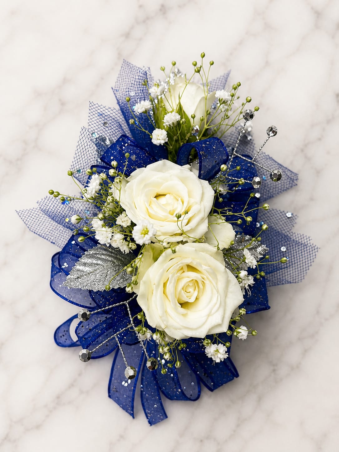 Moonlit Sapphire Formal Set - MAGNETIC BOUTONNIERE WARNING: CONTAINS STRONG MAGNETS. DO NOT USE IF YOU HAVE A PACEMAKER, DEFIBRILLATOR, INSULIN PUMP, COCHLEAR IMPLANT, NEUROSTIMULATOR, OR OTHER IMPLANTED MEDICAL DEVICE. KEEP AWAY FROM ELECTRONICS, CREDIT CARDS, HOTEL KEY CARDS, AND CHILDREN.  Moonlit Sapphire  White roses, sapphire blue ribbon, silver accents, rhinestone details, and delicate filler flowers come together in Moonlit Sapphire, an elegant corsage and boutonniere collection for prom, homecoming, dances, weddings, and formal events.  Standard includes a wrist corsage with 3 spray roses and a matching magnetic boutonniere with 2 roses. Deluxe includes 4 spray roses in the corsage. Premium includes 5 spray roses for additional fullness and flower coverage. All boutonnieres include 2 roses.  Perfect for prom flowers, wrist corsages, magnetic boutonnieres, corsage and boutonniere sets, homecoming flowers, dance flowers, and formal event flowers.  Because each corsage and boutonniere is individually designed, no two sets will be exactly alike. Ribbon, filler flowers, bracelets, decorative accents, and other design elements may vary based on availability. While individual details may differ, the overall color palette, roses and/or flowers, style, and appearance of the Moonlit Sapphire collection will be maintained.