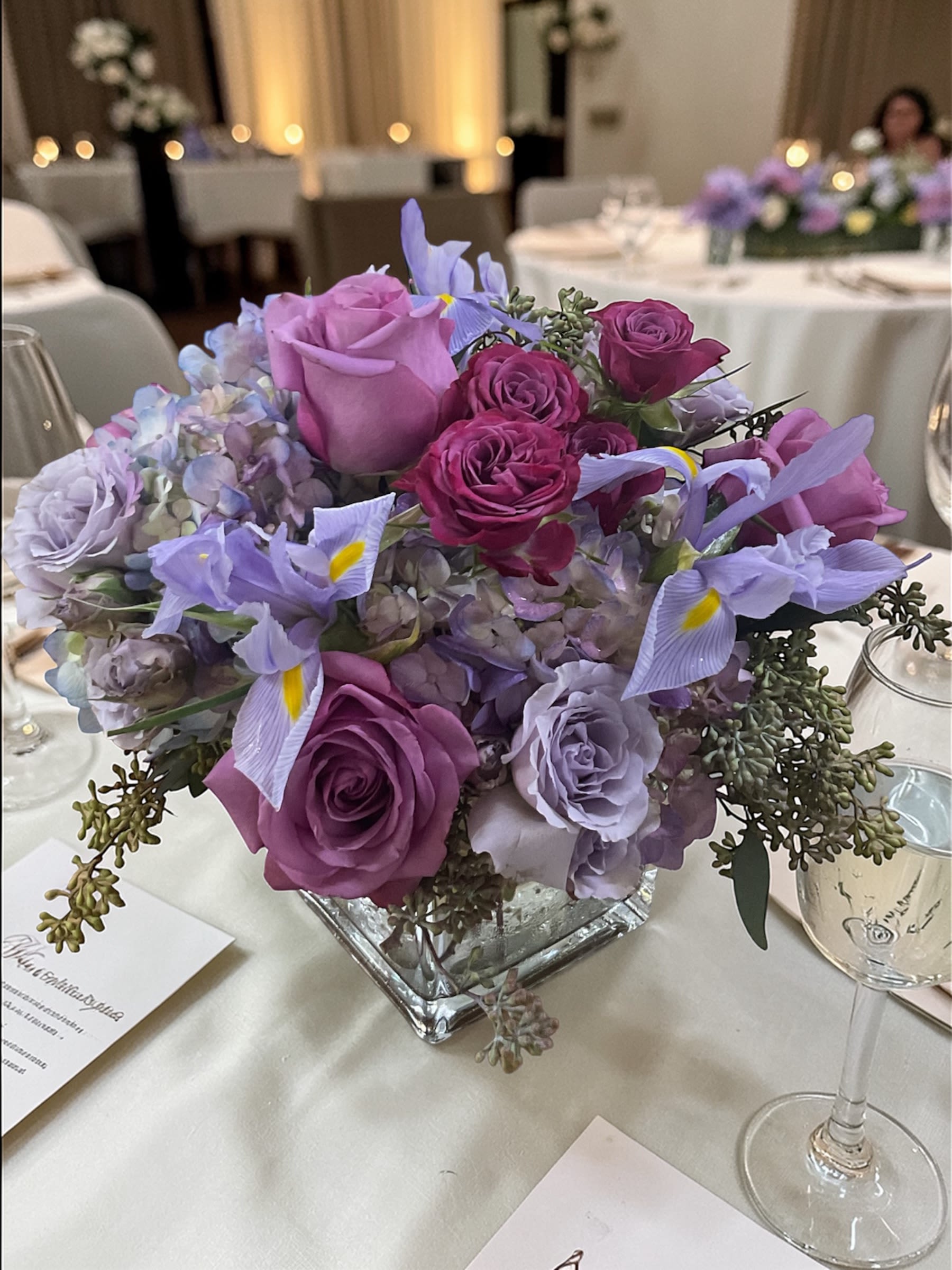 Lavender Skies Wedding Centerpiece  - Lavender Skies Wedding Centerpiece features lavender roses, lavender spray roses, hydrangeas, iris, and complementary greenery arranged in a romantic garden-inspired design. Soft shades of lavender, lilac, and periwinkle create an elegant wedding centerpiece filled with texture, color, and movement. Designed as part of the Lavender Skies wedding collection, this arrangement brings together classic wedding flowers and timeless styling to create a beautiful focal point for wedding receptions, sweetheart tables, head tables, and guest tables.  The combination of roses, spray roses, hydrangeas, and iris creates a lush, full appearance that adds beauty and sophistication to wedding décor while maintaining a soft, romantic feel. Whether used individually or throughout the reception space, these wedding centerpieces help create a cohesive floral design from ceremony to reception. Popular for couples searching for lavender wedding flowers, romantic wedding centerpieces, hydrangea centerpieces, and garden-inspired wedding décor, this design offers classic beauty with a fresh, modern touch.  Flowers and specific varieties may vary based on seasonal and market availability. Substitutions may be necessary; however, the overall style, color palette, quality, value, and feel of the design will be maintained as closely as possible.