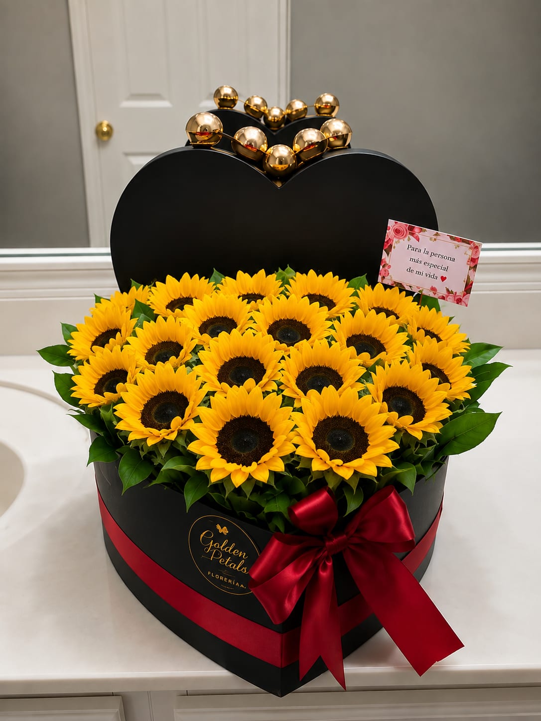 Heart-shaped box of yellow sunflowers with a red ribbon and gold topper