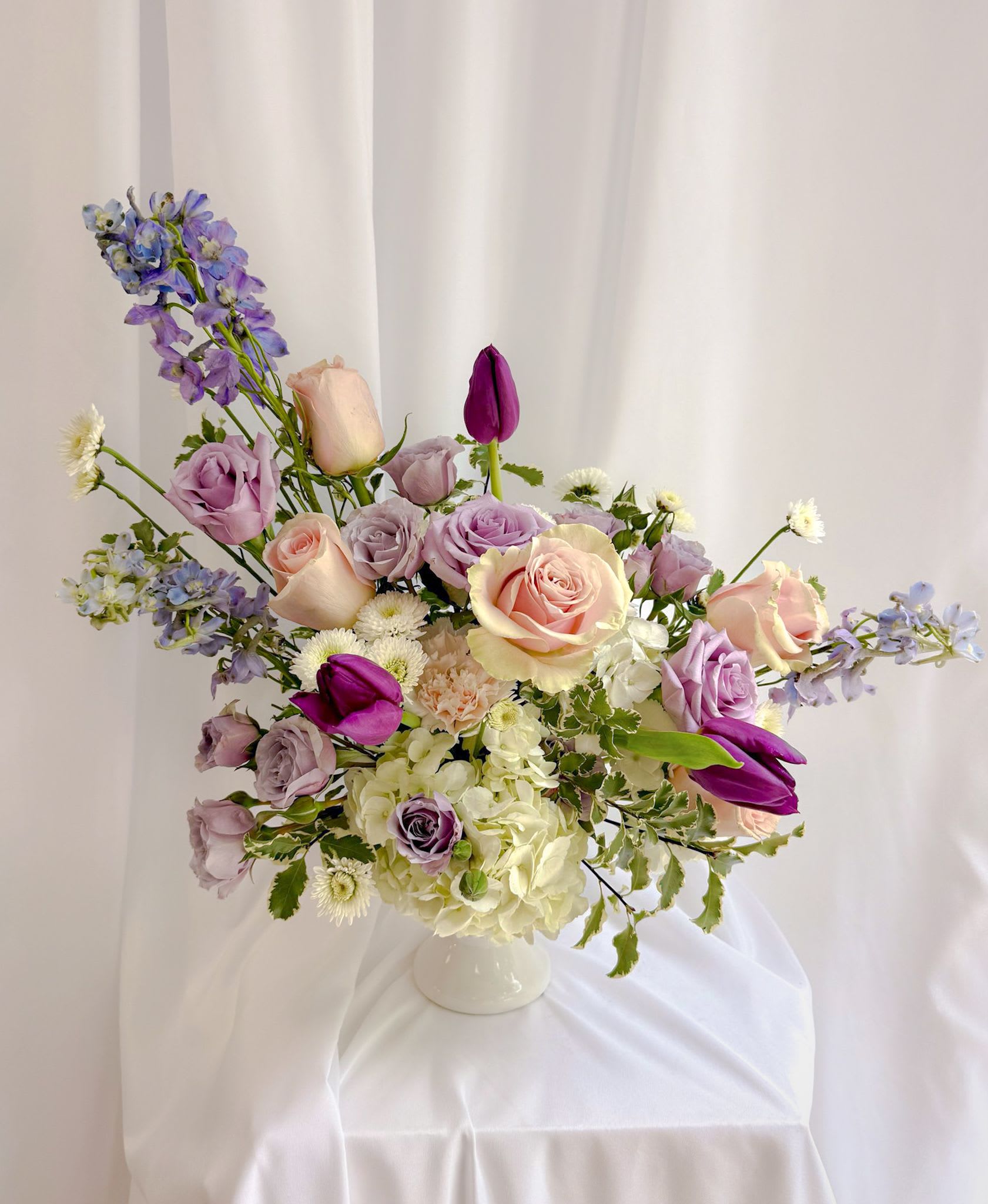 Blushing Garden - A soft, elegant mix of blush roses, lavender blooms, creamy hydrangea, and vibrant tulips in a classic white vase. Light, romantic, and perfect for celebrating Mom with beauty and grace.