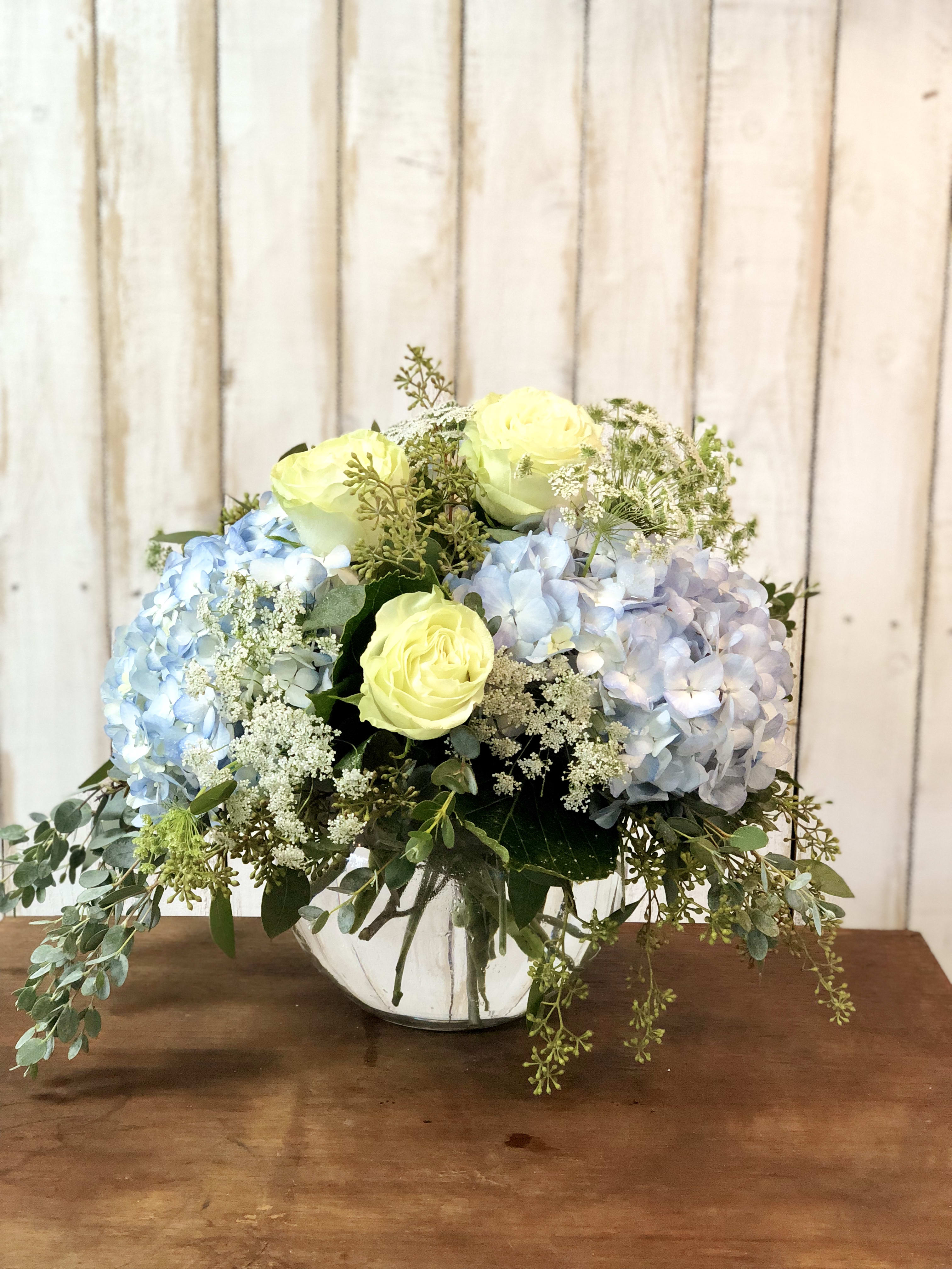 Blue and lavender hydrangeas with pale yellow roses in a glass vase