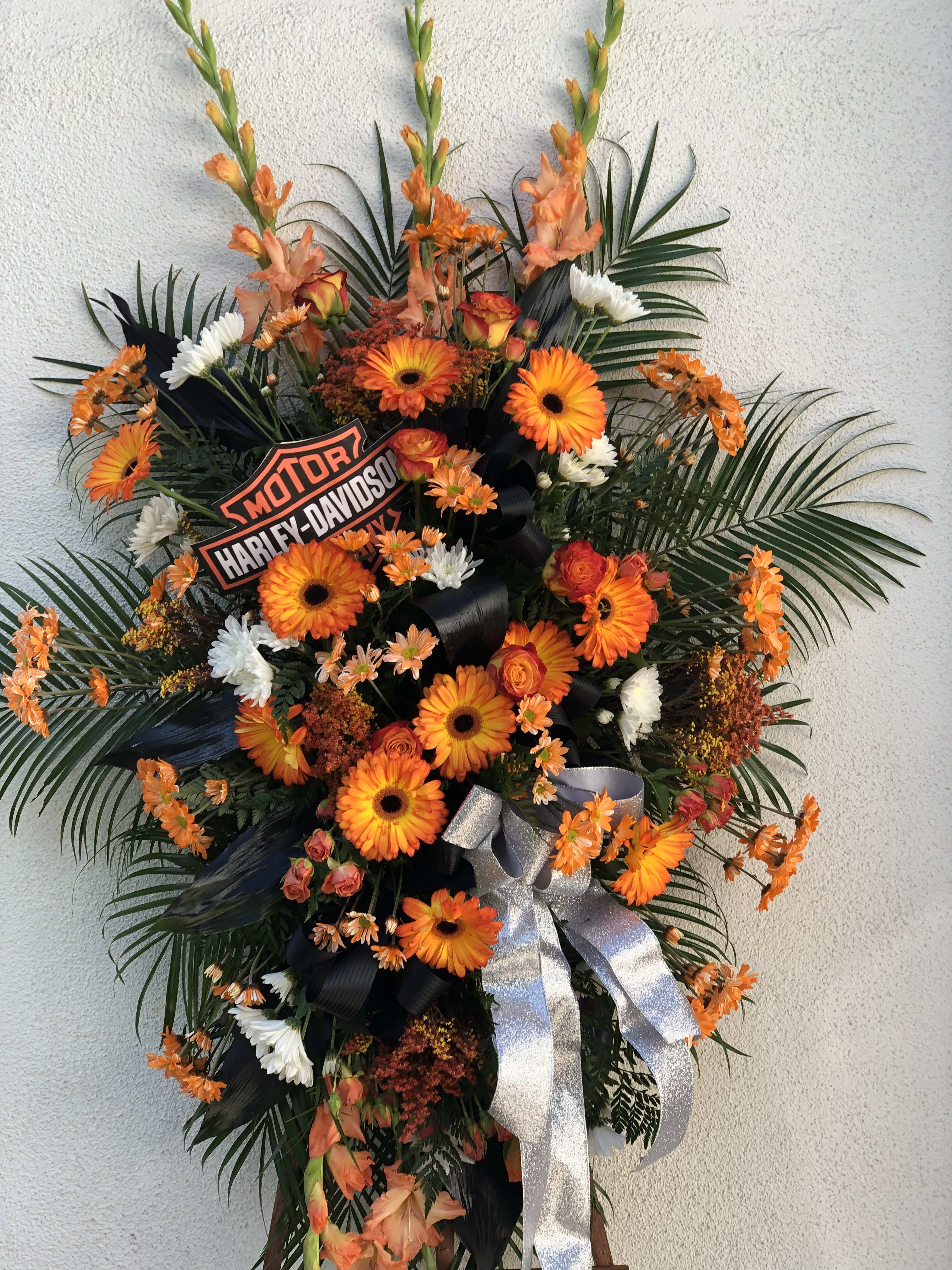 Tall standing spray with orange gerbera daisies, white flowers, palm fronds, and a Harley-Davidson logo sign.