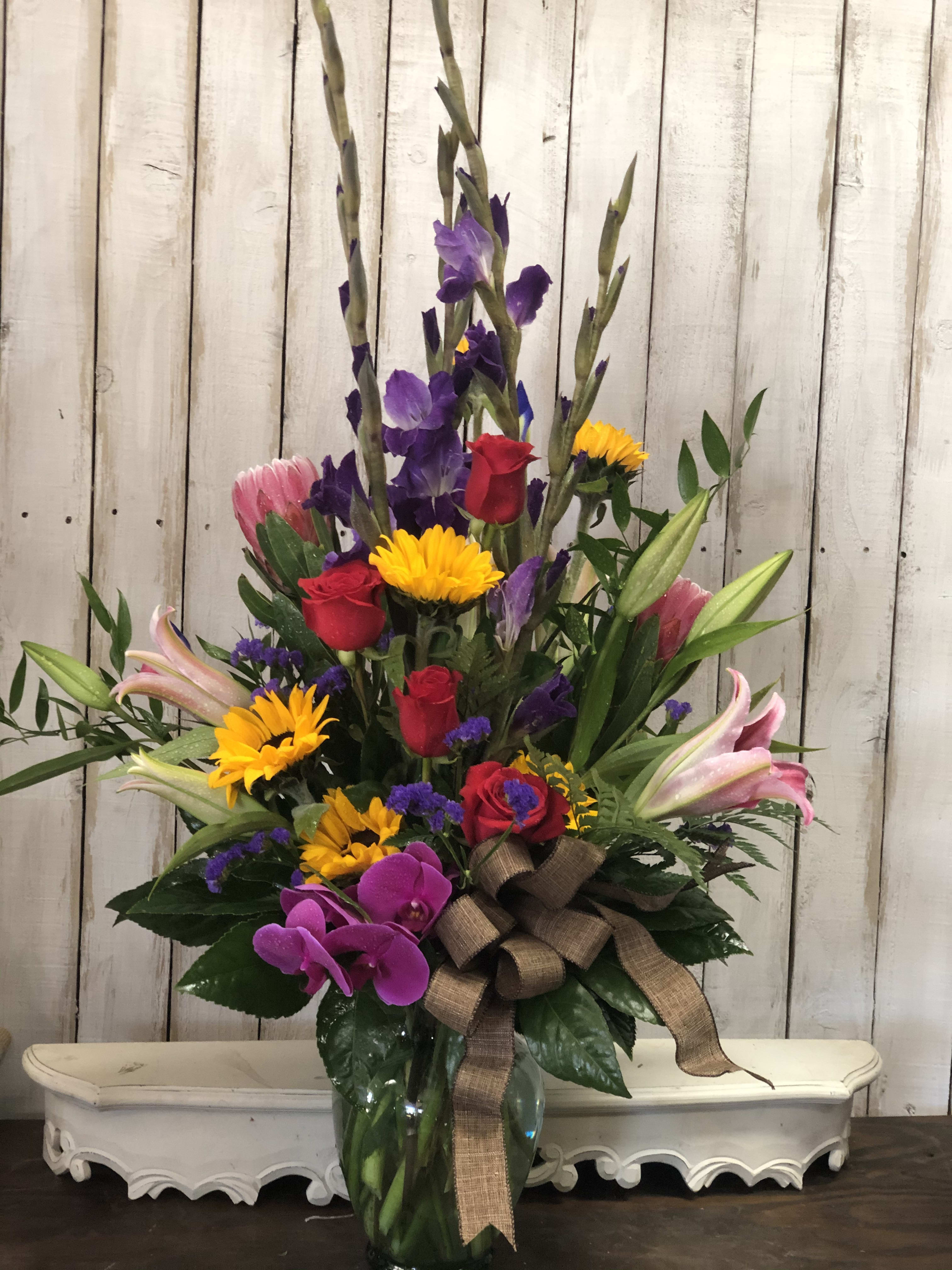 Tall mixed bouquet in a glass vase with roses, lilies, orchids, and yellow daisies