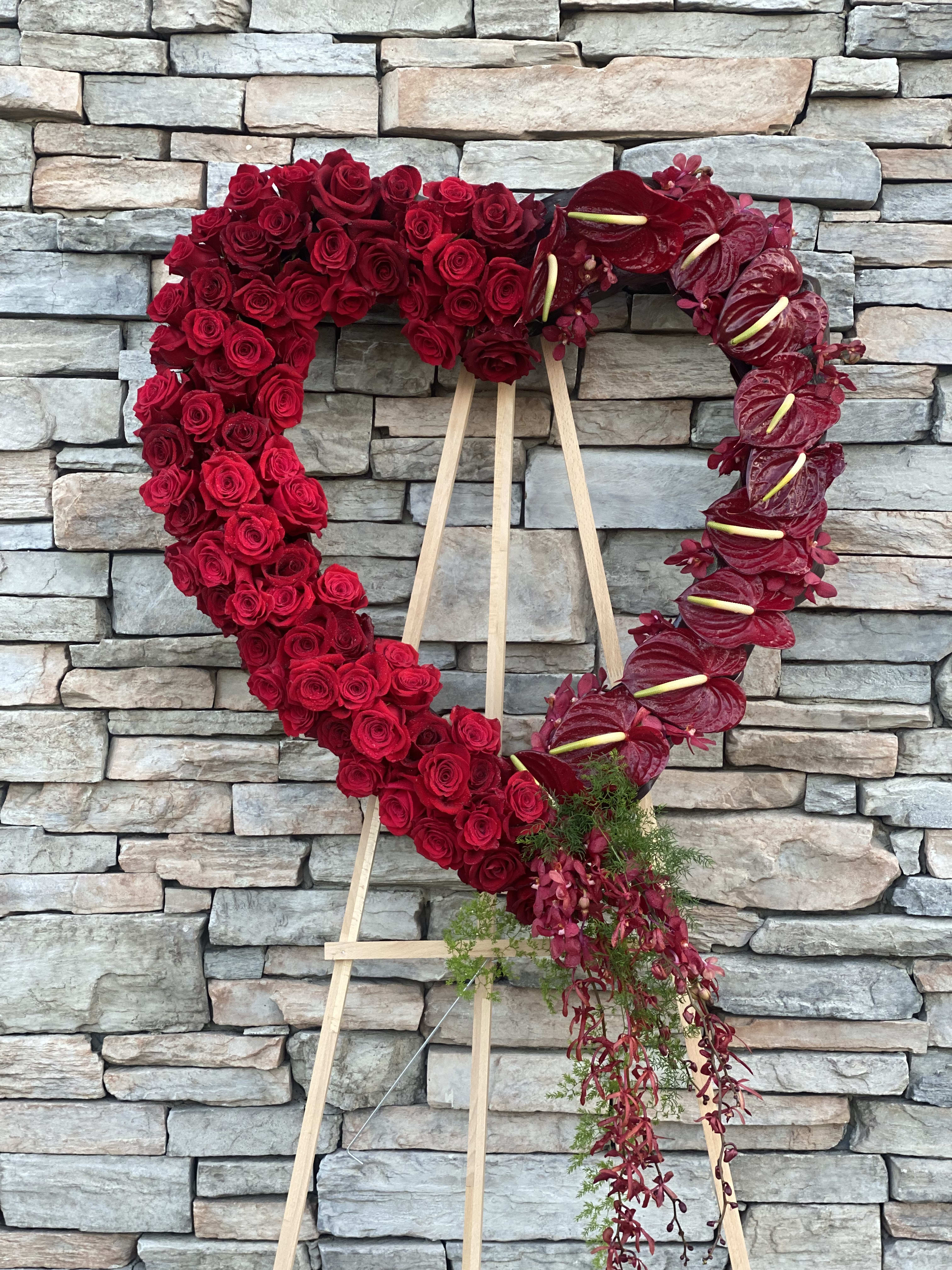 Open Heart Red Rose Tribute  - Honor a life beautifully remembered with our Open Heart Red Rose Tribute, a stunning and meaningful expression of love, remembrance, and deep sympathy. Designed in the shape of an open heart, this elegant standing spray symbolizes everlasting love and a heartfelt farewell.  This custom design features rich red roses, striking red anthuriums, and lush tropical foliage, thoughtfully arranged with flowing orchid accents that cascade gracefully from the base. The combination of textures and deep red tones creates a bold yet graceful tribute, offering comfort and honoring a loved one in a truly unforgettable way.  Each piece is handcrafted with care and intention by our floral designers, ensuring every arrangement reflects both beauty and meaning. This design is ideal for funeral services, memorials, or celebration of life ceremonies.  Available for delivery in Rancho Cucamonga and throughout the Inland Empire, we will ensure your tribute arrives with care, dignity, and respect.