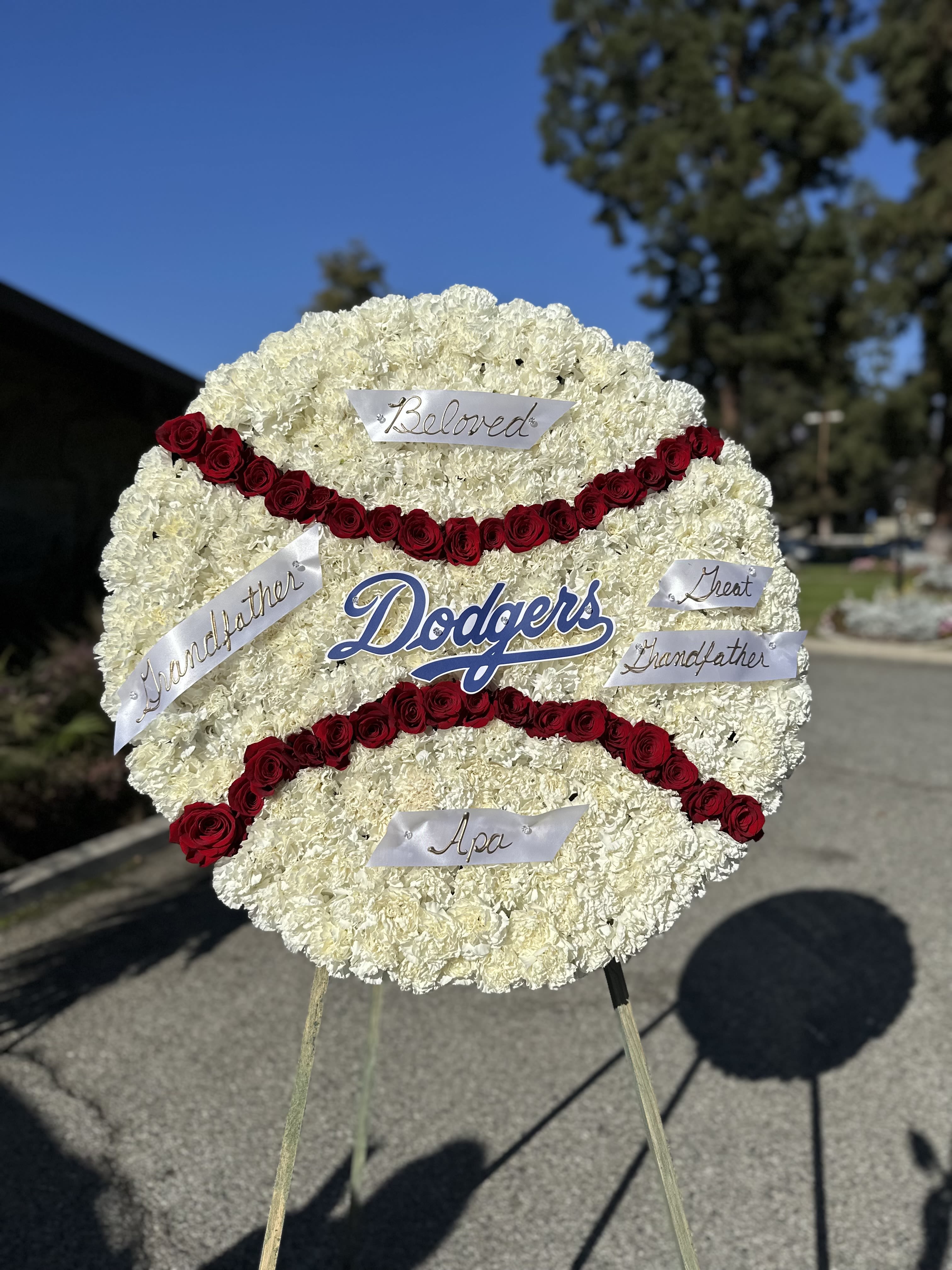 Dodger fan  - Celebrate the life of a devoted Dodgers fan with a stunning custom floral standing arrangement inspired by the iconic Dodgers baseball. Handcrafted with care, this design features a lush bed of pristine white carnations, accented by striking red roses to recreate and symbolize the classic baseball stitching. Perfect for memorials or celebrations of life, this unique piece reflects a love for the game and the cherished memories it holds.