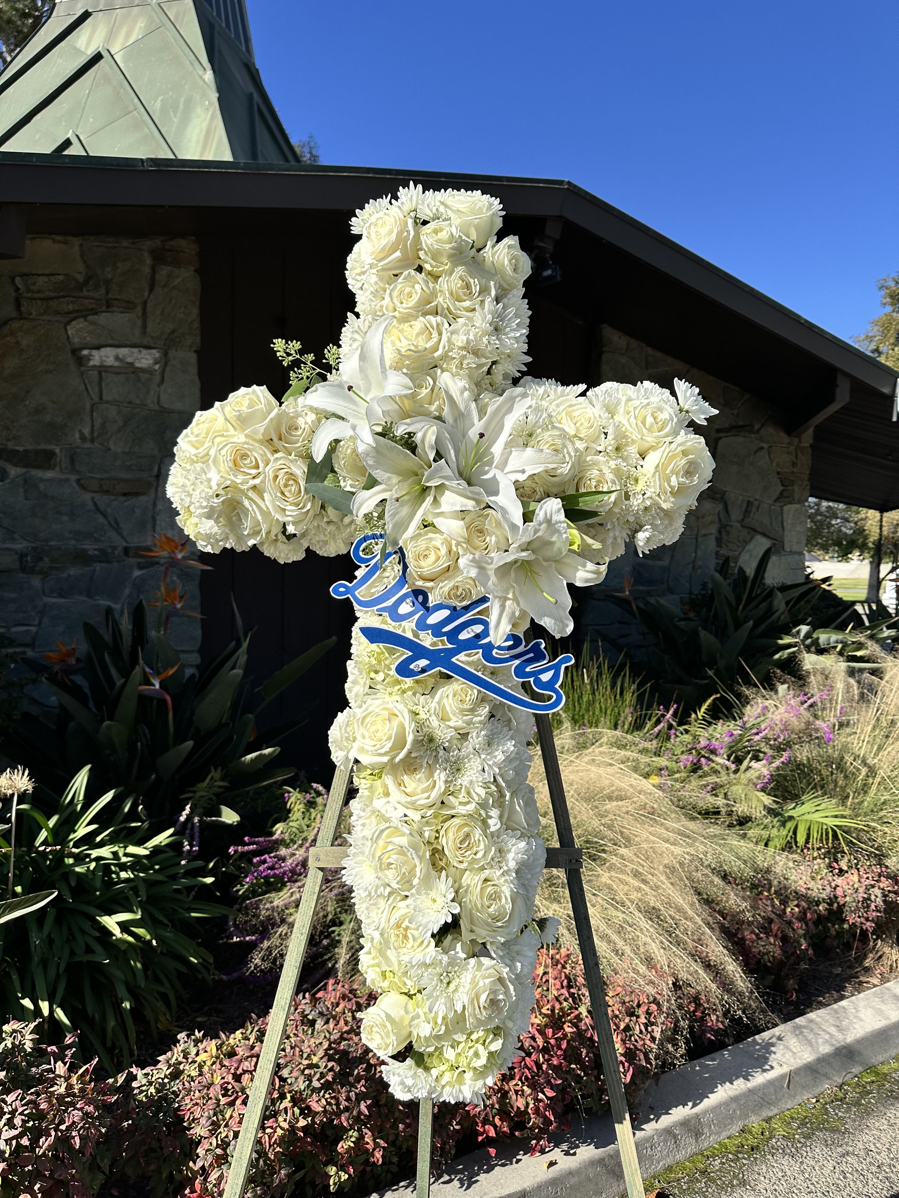 Rest in peace, dodger fan  - This elegant 36-inch cross floral arrangement is a heartfelt tribute to honor a loved one's faith and passions. Featuring a stunning blend of three dozen white roses, Casa Blanca lilies, carnations, and chrysanthemums, this design exudes grace and serenity. At its center, a custom Dodgers logo adds a personal touch, celebrating the legacy of a true fan. Perfect for memorial services or celebrations of life, this arrangement beautifully combines devotion, love, and team spirit.