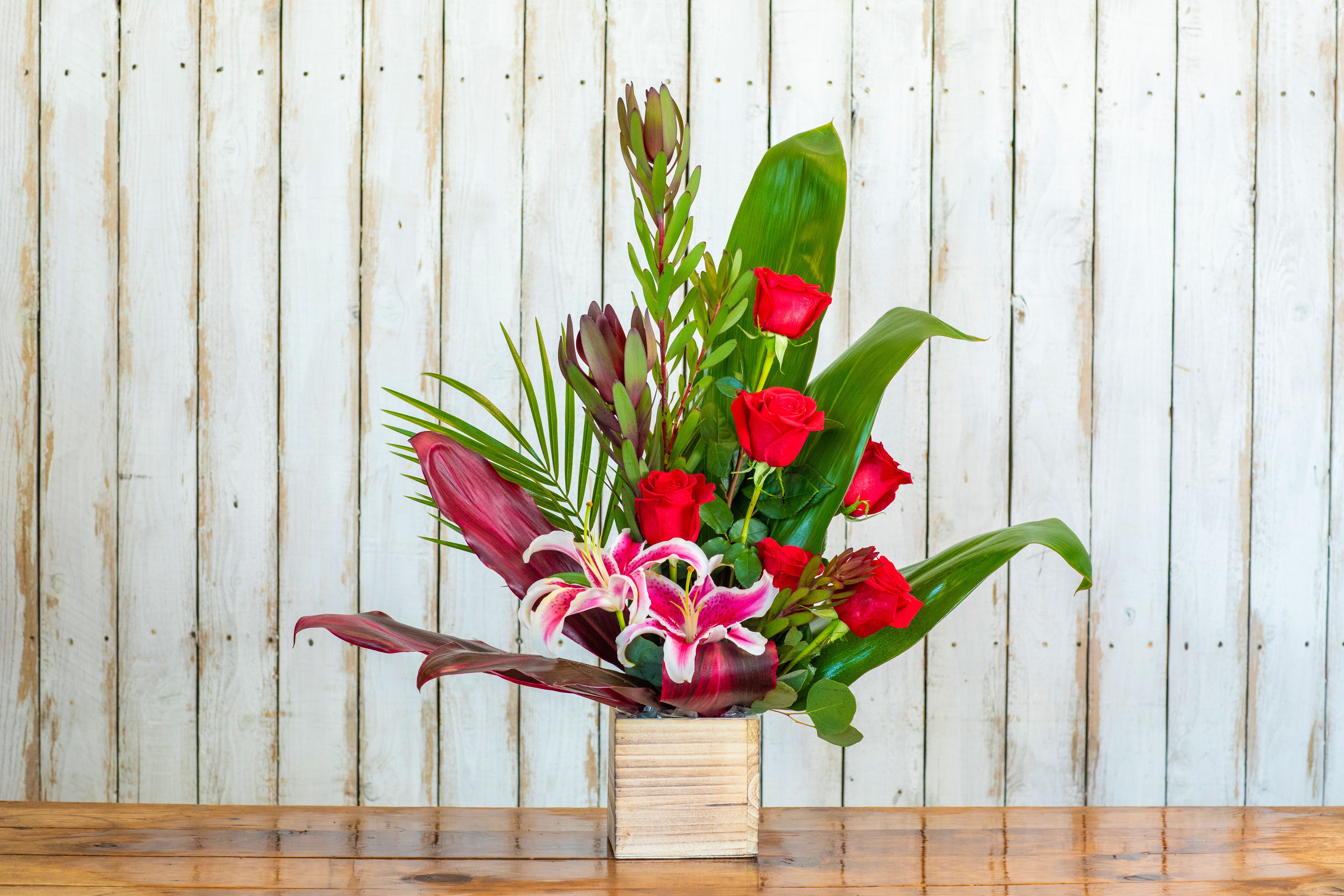 Modern Love Tropical Mix - Make a bold statement with the Modern Love Tropical Mix, a dramatic and contemporary arrangement designed by your creative Rancho Cucamonga Florist. This unique piece blends passionate classic romance with exotic, modern flair.  The design features brilliant red roses—symbols of deep love—paired with vibrant pink stargazer lilies. These beautiful blooms are accented by striking tropical elements, including long green and burgundy leaves, palm fronds, and a distinct red-tipped protea. This vertical arrangement is nestled in a stylish, light wood cube container, enhancing its modern aesthetic.  As the premier Rancho Cucamonga flower shop, we craft arrangements that stand out. This arrangement is an exceptional choice for a passionate anniversary gift, a chic birthday bouquet, or any occasion where you want to send a high-fashion, high-impact message. It's the perfect way to show profound emotion with an artistic touch.  We guarantee the freshness of every bloom and offer reliable same-day flower delivery across the entire Rancho Cucamonga area. Trust our expert florists to deliver this spectacular, one-of-a-kind arrangement with care. Order the Modern Love Tropical Mix online today from your dedicated local Rancho Cucamonga Florist!