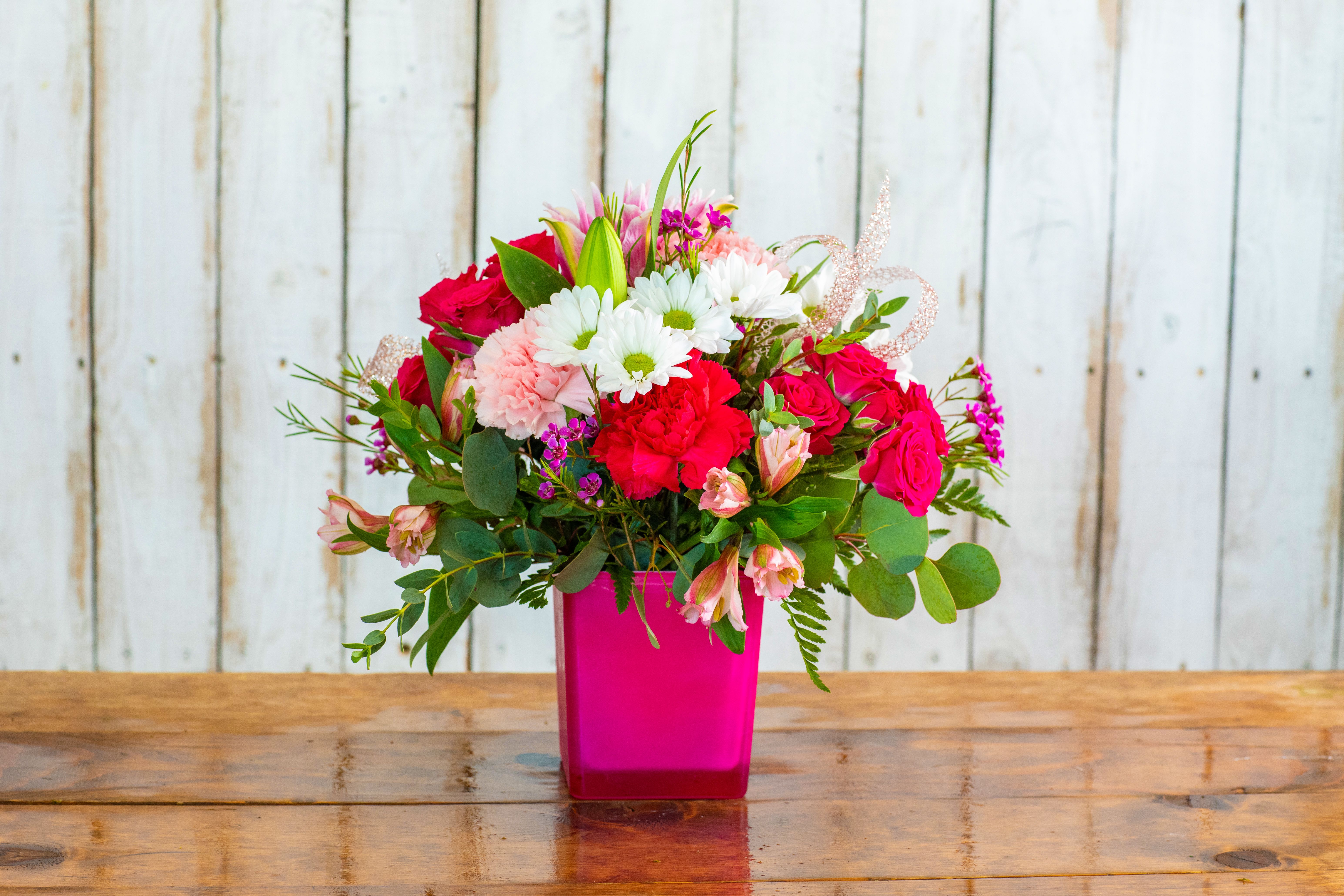 Fuchsia Flirtation Bloom - Bold, vibrant, and full of life, our Fuchsia Flirtation Bloom is designed to make a lasting impression. This eye-catching arrangement features rich red roses, hot pink spray roses, cheerful white daisies, and lush seasonal blooms, all beautifully accented with fresh greenery.  Designed in a striking fuchsia glass vase, this arrangement is perfect for birthdays, anniversaries, romantic surprises, or any moment that calls for something bright, fun, and unforgettable.  Each piece is thoughtfully handcrafted by our floral designers, giving it a fresh, garden-inspired feel with a modern touch. No two arrangements are exactly alike—making your gift truly one-of-a-kind.  Available for same-day flower delivery in Rancho Cucamonga and throughout the Inland Empire, this vibrant bouquet is the perfect way to celebrate, surprise, and show appreciation in a big, beautiful way. 