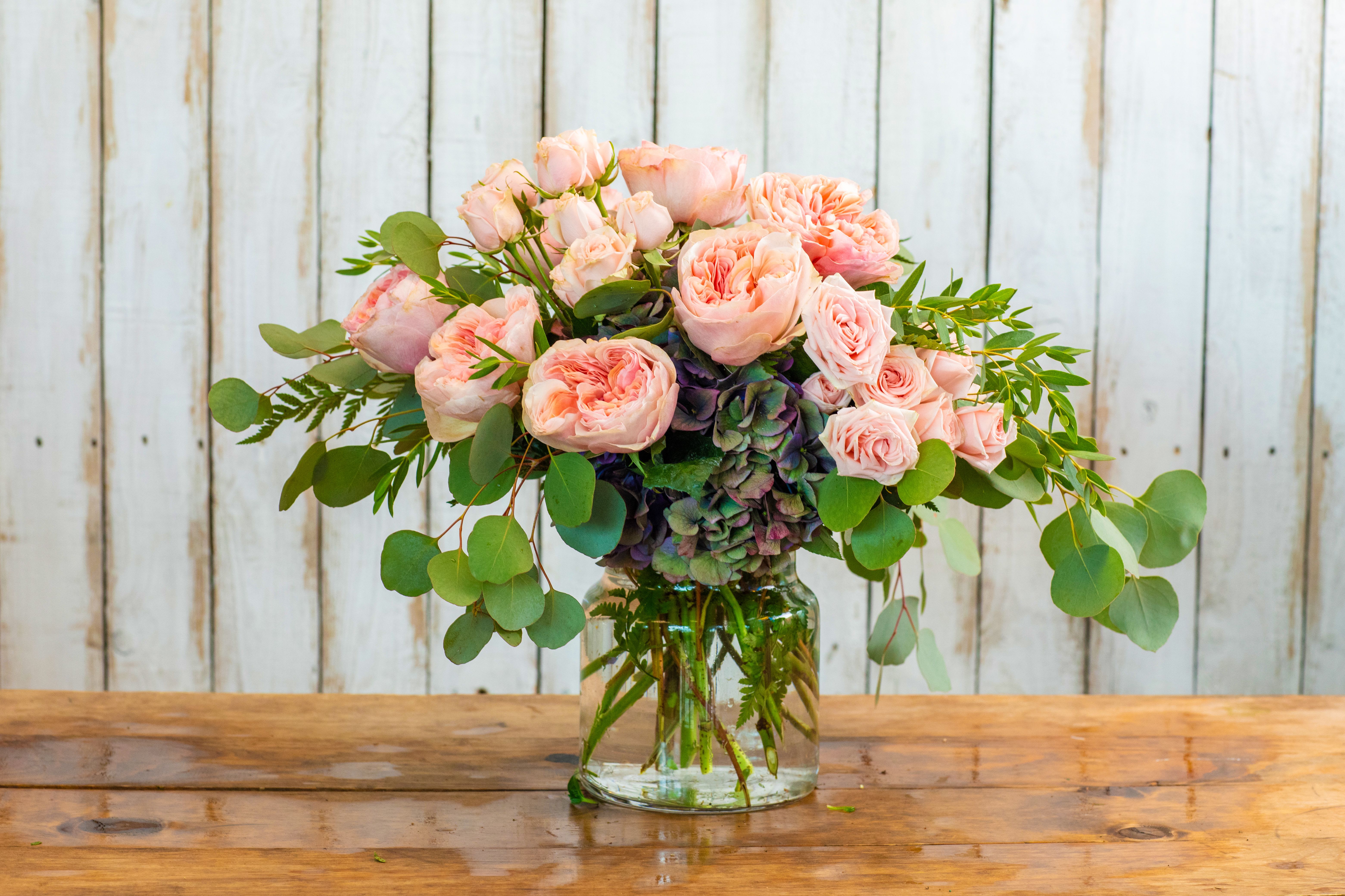Blushing Garden Romance - Indulge in pure elegance with the Blushing Garden Romance, a luxurious and enchanting arrangement lovingly crafted by your premier Rancho Cucamonga Florist. This exquisite bouquet is a celebration of soft textures and romantic hues.  It features an abundance of fragrant, full-blooming pink garden roses and delicate spray roses, perfectly nestled atop a base of deep purple and green hydrangeas. Lush, rounded eucalyptus leaves provide a beautiful, verdant frame. All these opulent blooms are artfully arranged in a classic clear glass vase, making it a sophisticated gift to send instantly.  As the leading Rancho Cucamonga flower shop, we make it easy to shop online for flowers that express profound affection. This breathtaking arrangement is the quintessential choice for a romantic anniversary gift, a heartfelt Mother's Day flowers surprise, a celebratory birthday bouquet, or a grand gesture to show your love.  We offer reliable same-day flower delivery throughout the entire Rancho Cucamonga area. Don't delay—order flowers now and trust our expertise to deliver a stunning and unforgettable arrangement. Buy flowers online today with confidence from your dependable local Rancho Cucamonga Florist!