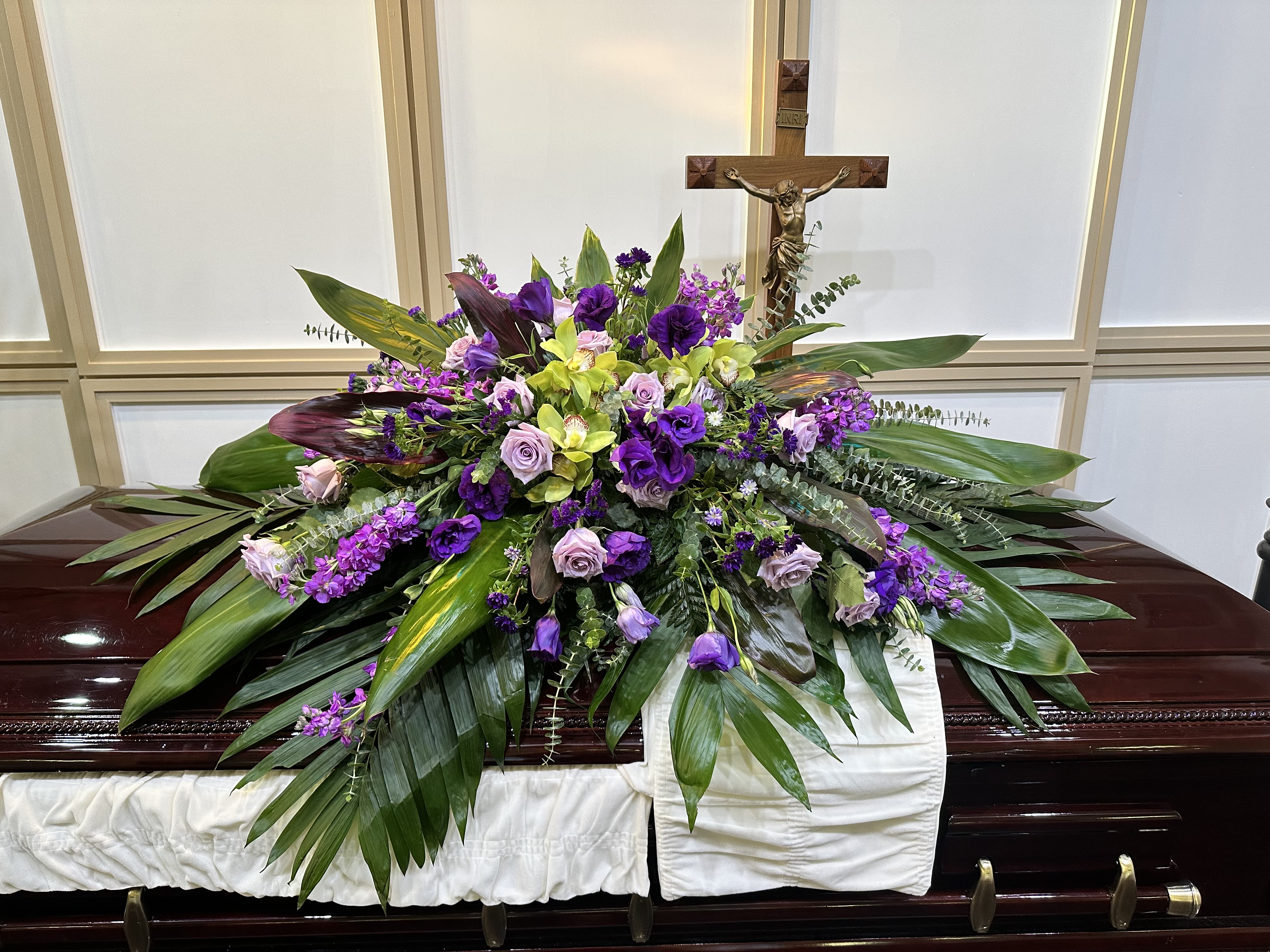 Purple and green floral casket spray with a crucifix behind it