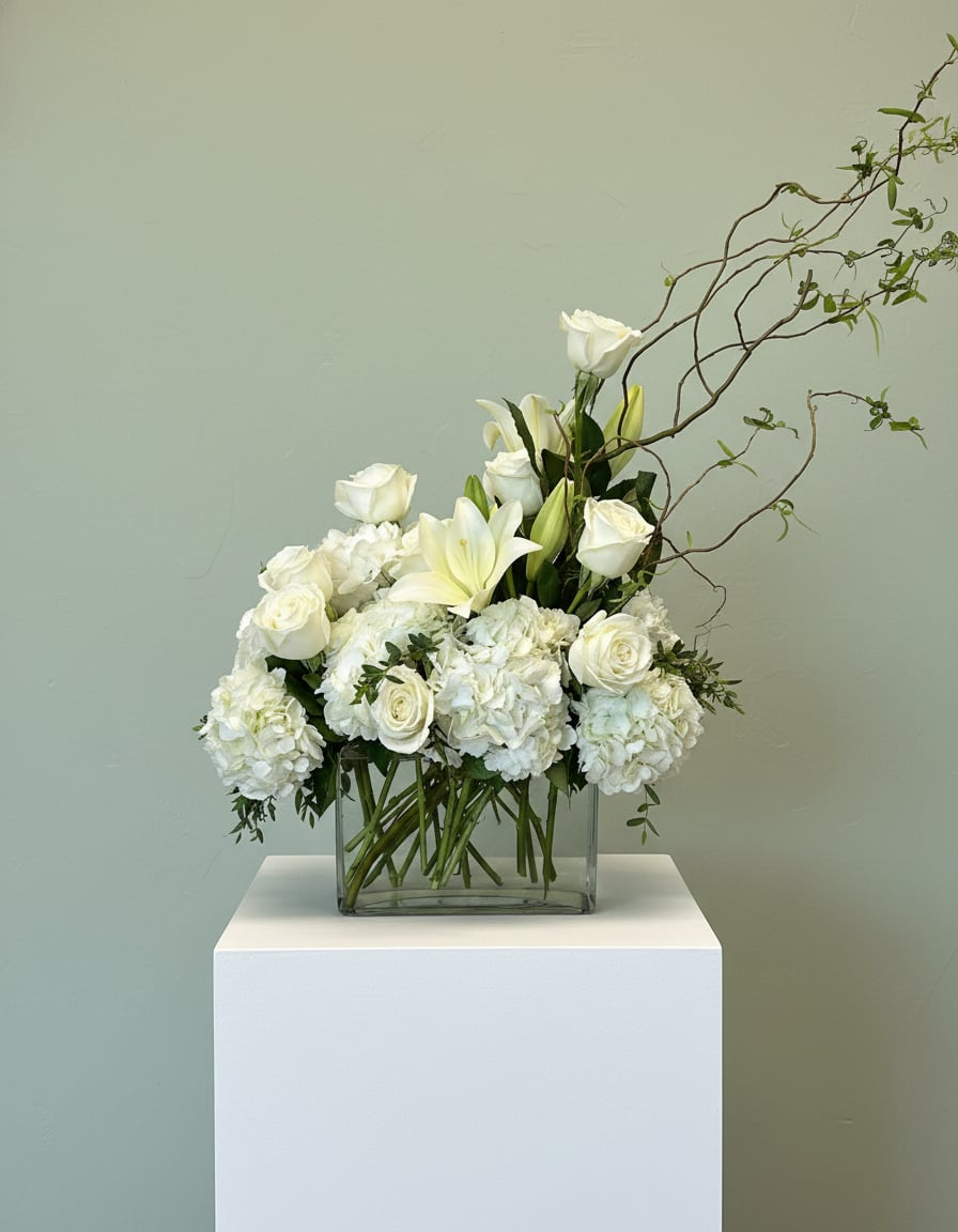 Heavenly Grace White Sympathy Arrangement - Honor a life beautifully remembered with our Heavenly Grace White Sympathy Arrangement, a timeless design created to express love, peace, and heartfelt condolences. This elegant arrangement features premium white roses, hydrangeas, and lilies, artfully designed in a modern glass vase with natural flowing branches for a soft, graceful touch. The all-white palette symbolizes purity, remembrance, and eternal love making it a perfect choice for funerals, memorial services, or sending comfort to grieving loved ones. Designed with a balance of lush fullness and airy movement, this arrangement offers both sophistication and serenity. Handcrafted with care by our expert designers at Tommy Austin Florist, this piece is ideal for delivery to homes, funeral services, churches, and mortuaries.