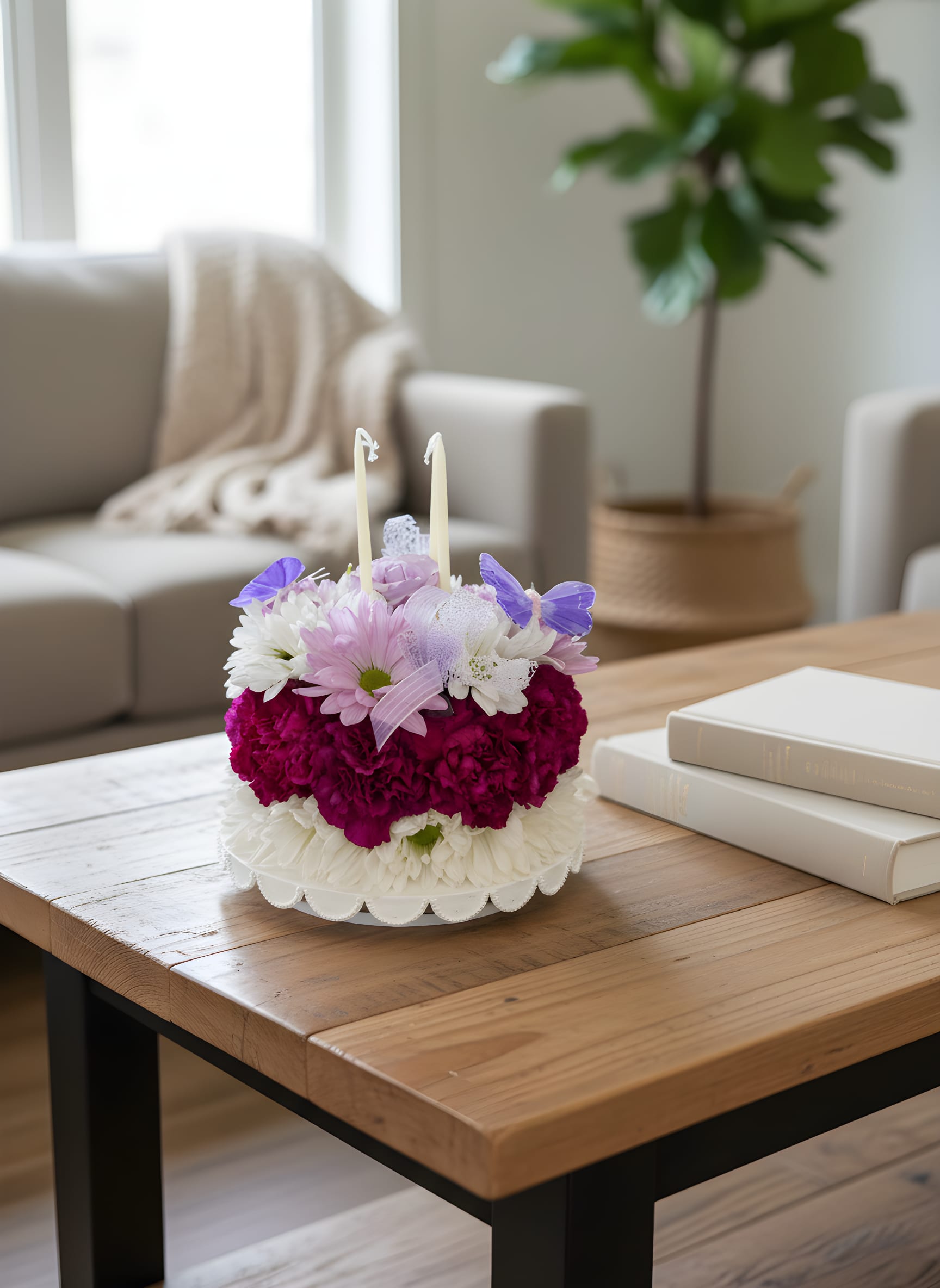 Butterfly Birthday Bloom Cake - Celebrate someone’s special day with a gift they’ll never forget! Our Butterfly Birthday Bloom Cake is a handcrafted floral arrangement designed to look like a beautiful birthday cake. Created with fresh seasonal flowers in cheerful shades of pink, white, and lavender, it features elegant butterflies, decorative ribbon, two birthday candles, and a reusable cake stand.  Perfect for birthdays of all ages, this unique floral gift is a wonderful alternative to traditional birthday presents and is sure to become the centerpiece of any celebration.  Includes: * Fresh seasonal flowers * Decorative butterflies * Two birthday candles * Reusable cake stand  Please Note: This is a floral arrangement designed to resemble a birthday cake. It is NOT edible. Flowers and butterfly accents may vary slightly based on seasonal availability while maintaining the overall style and color palette.