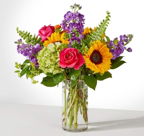 Mixed bouquet of sunflowers, pink roses, and purple blooms in a glass vase