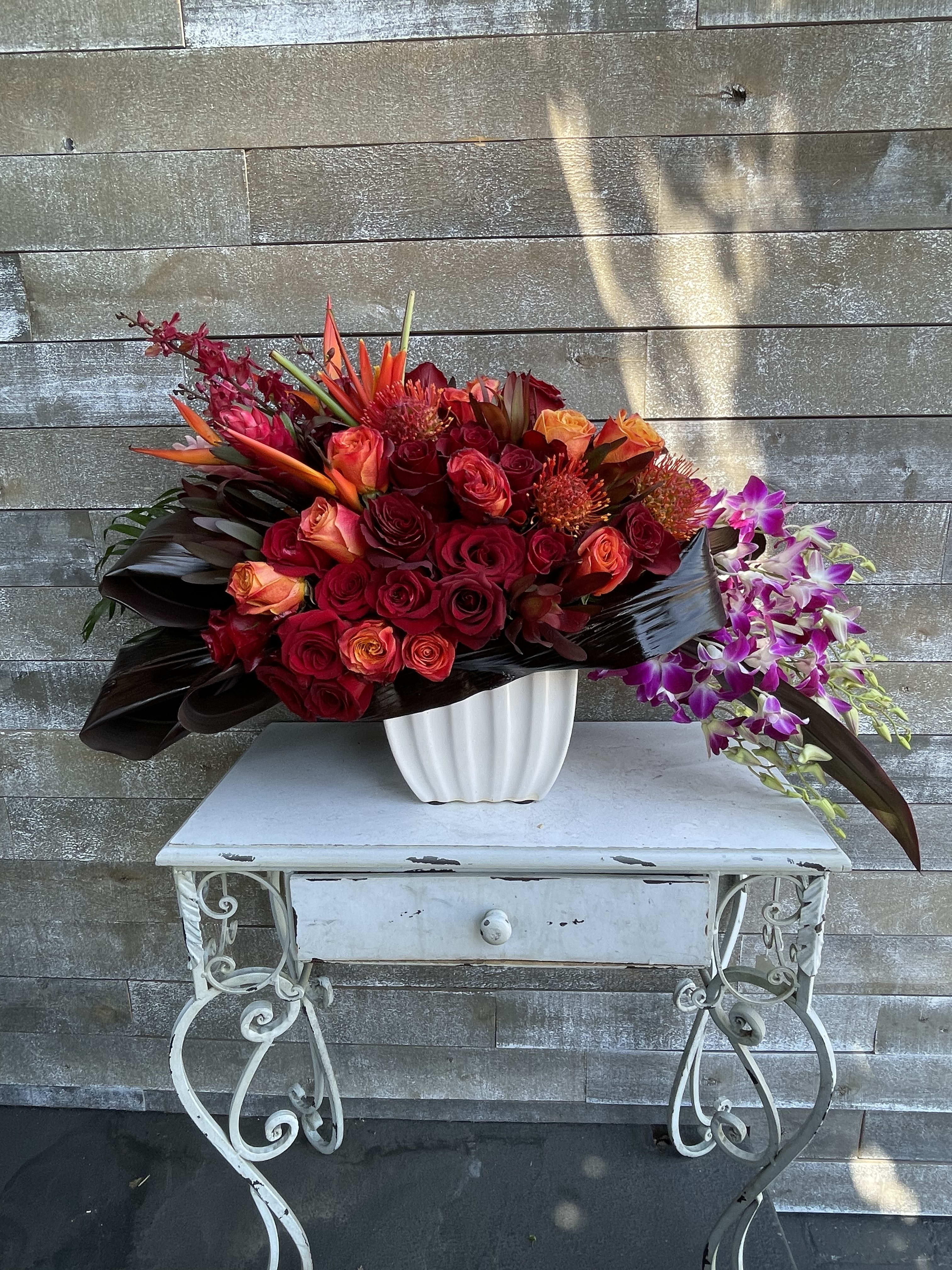 Large red and orange floral arrangement in a white vase with purple orchids