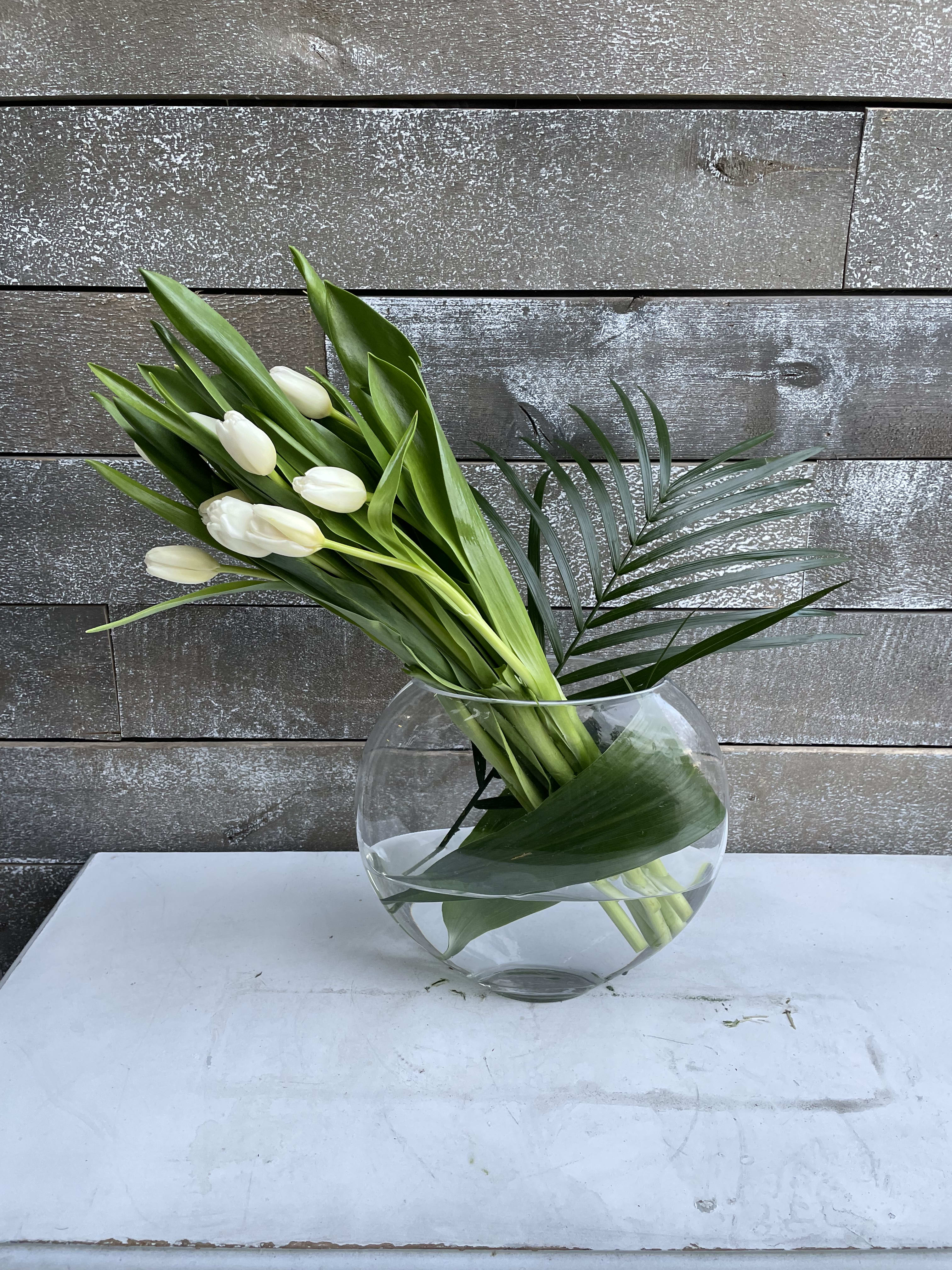 White tulips arranged in a round glass vase with palm leaves