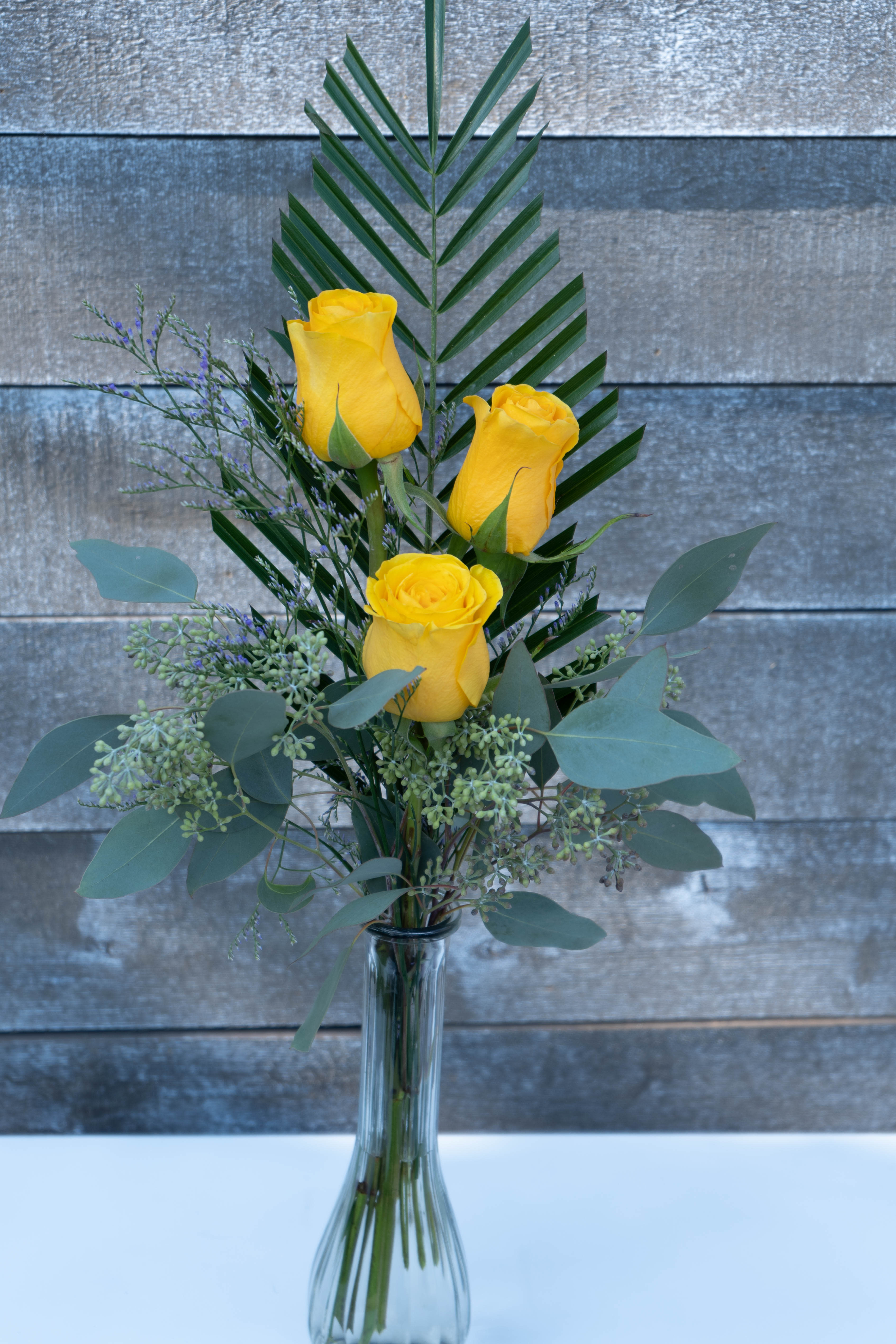 Three yellow roses in a clear glass vase with mixed greenery