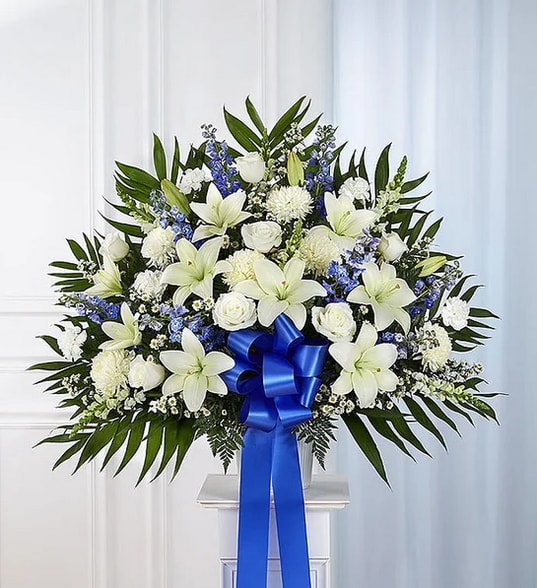 White lilies and roses with blue flowers and a ribbon on a stand