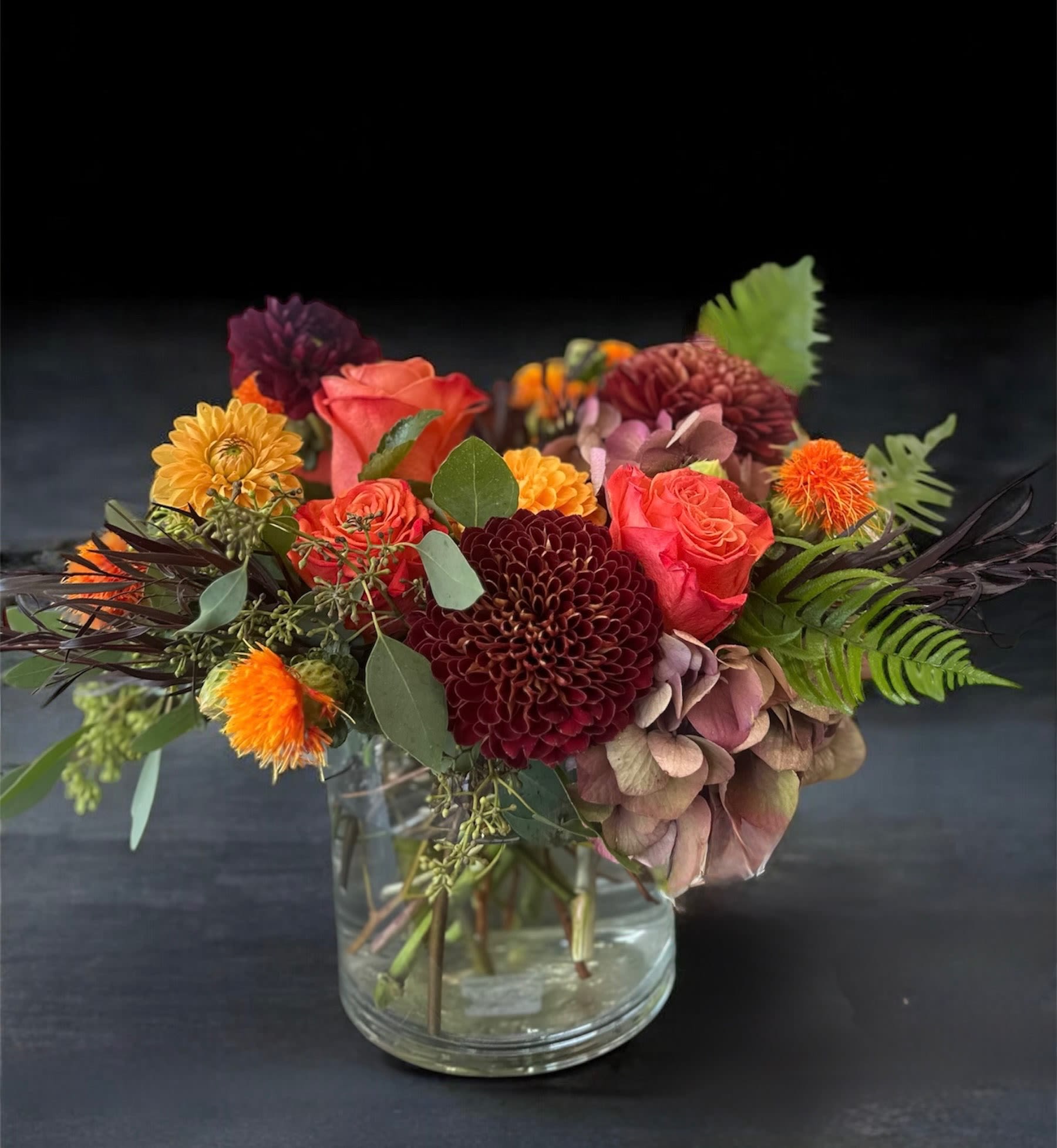 Mixed bouquet of orange, burgundy, and pink flowers in a clear glass vase