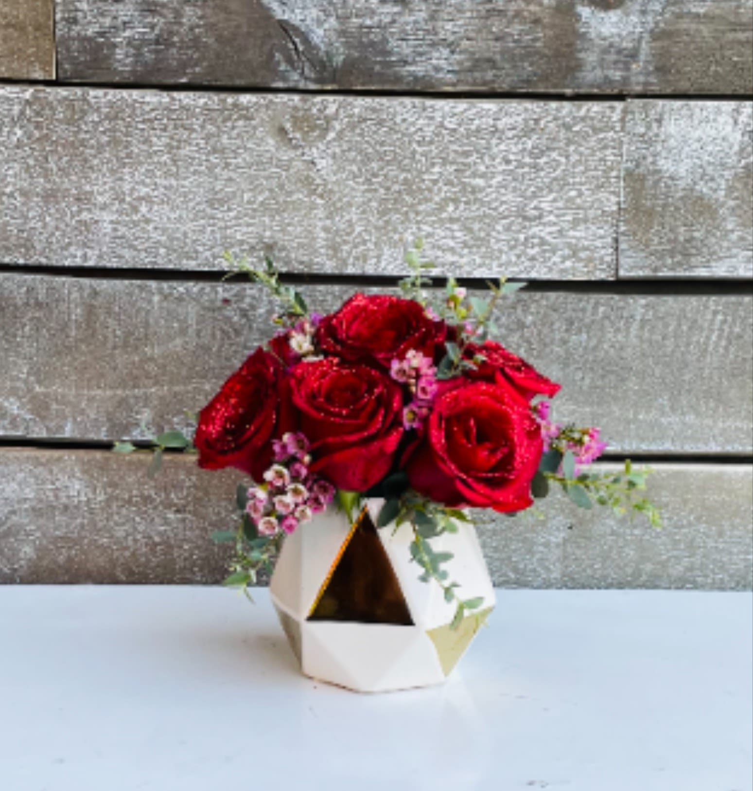 Red roses arranged in a geometric white vase with small pink filler flowers