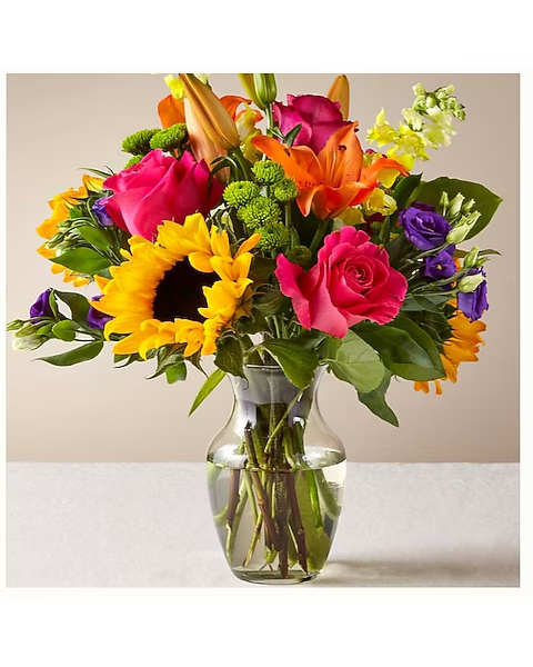 Colorful mixed bouquet in a clear glass vase with sunflowers, roses, lilies, and purple blooms