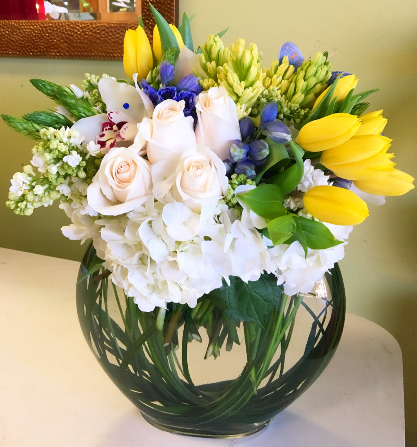 Bouquet of white roses, yellow tulips, and blue flowers in a round glass vase