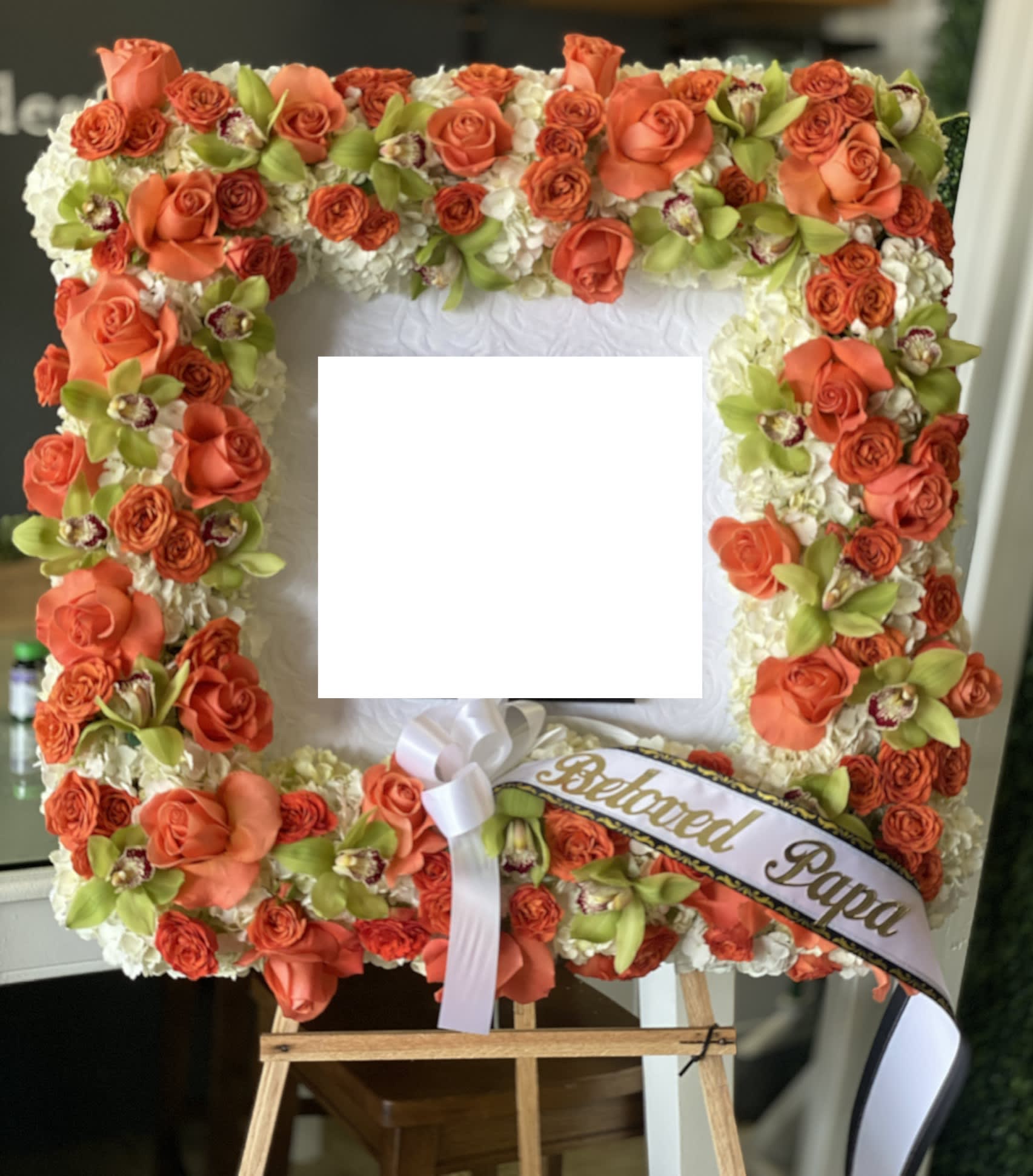 Square floral tribute with orange roses and white flowers on an easel