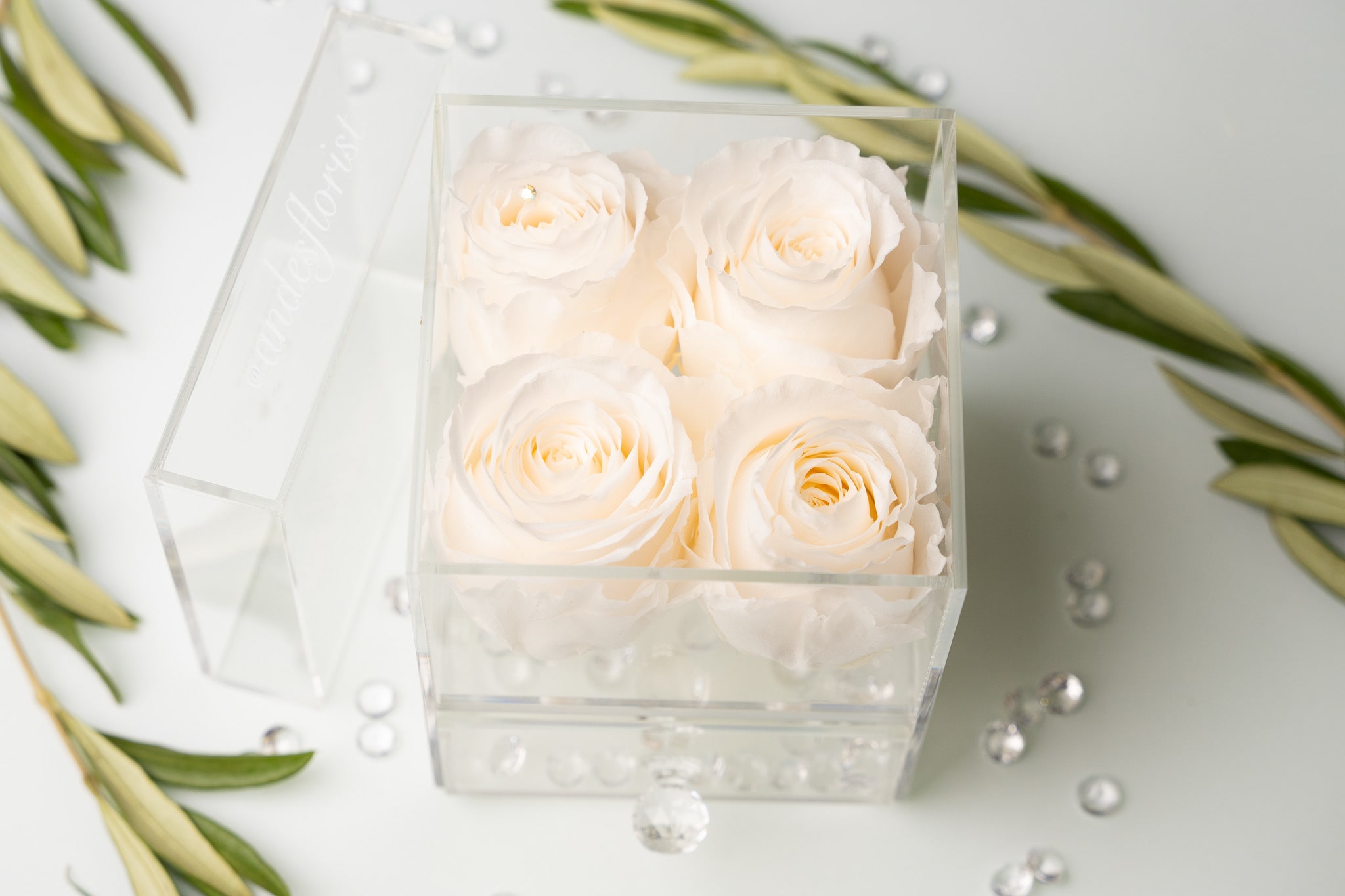 Four white roses in a clear acrylic box with scattered crystals