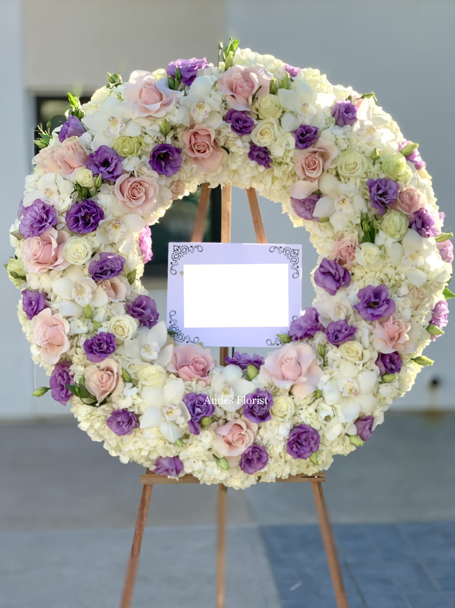 Large floral wreath of pink, purple, and white blooms on an easel with a blank sign