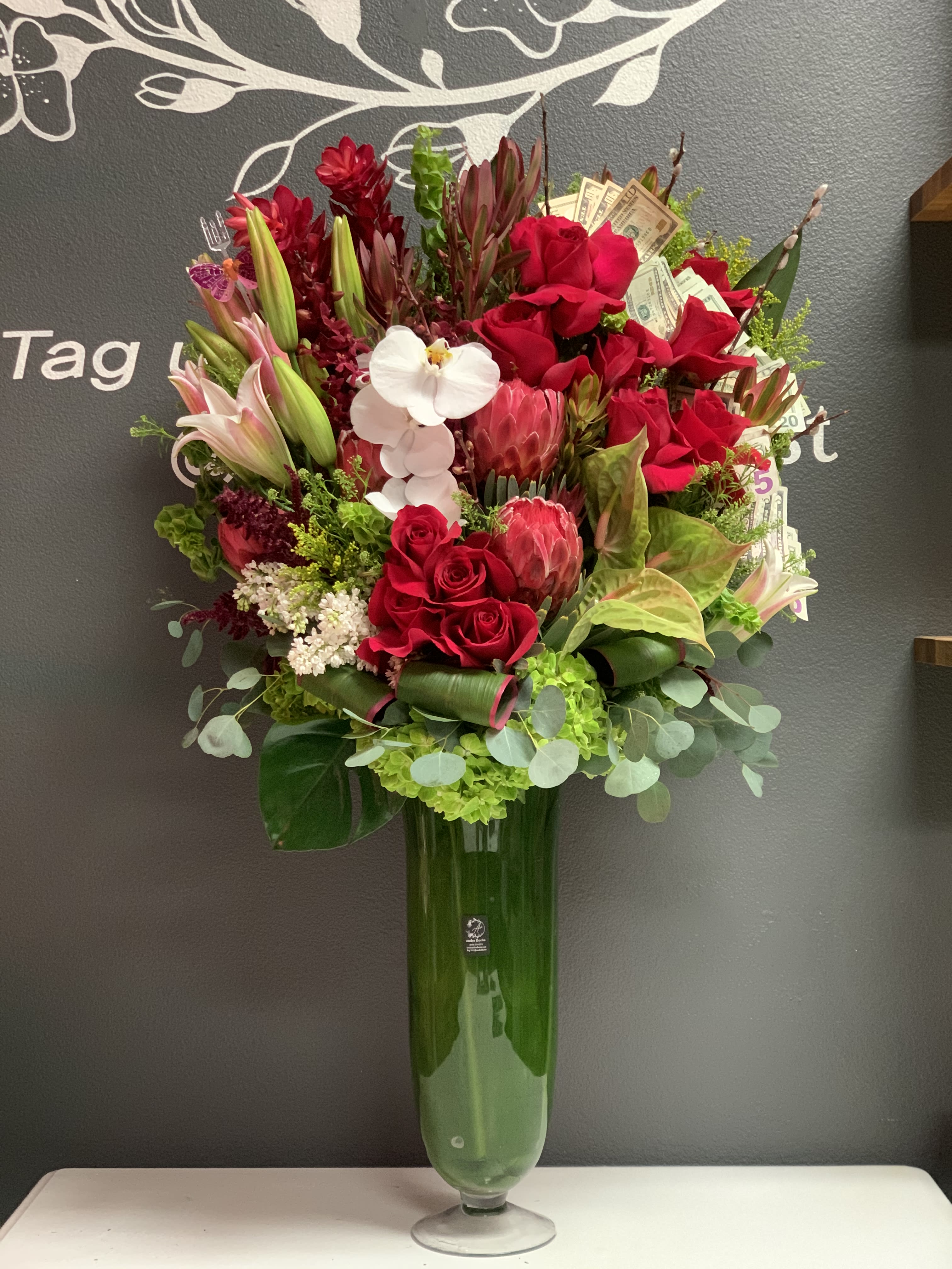 Tall red floral arrangement in a glass vase with a white orchid and dollar bills