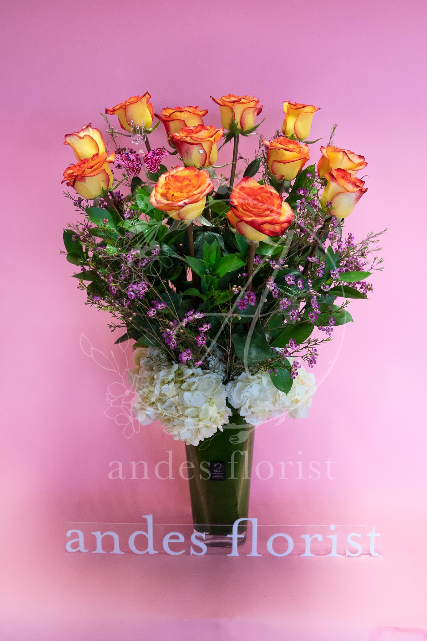 Orange-yellow roses in a tall vase with white hydrangeas and purple filler flowers