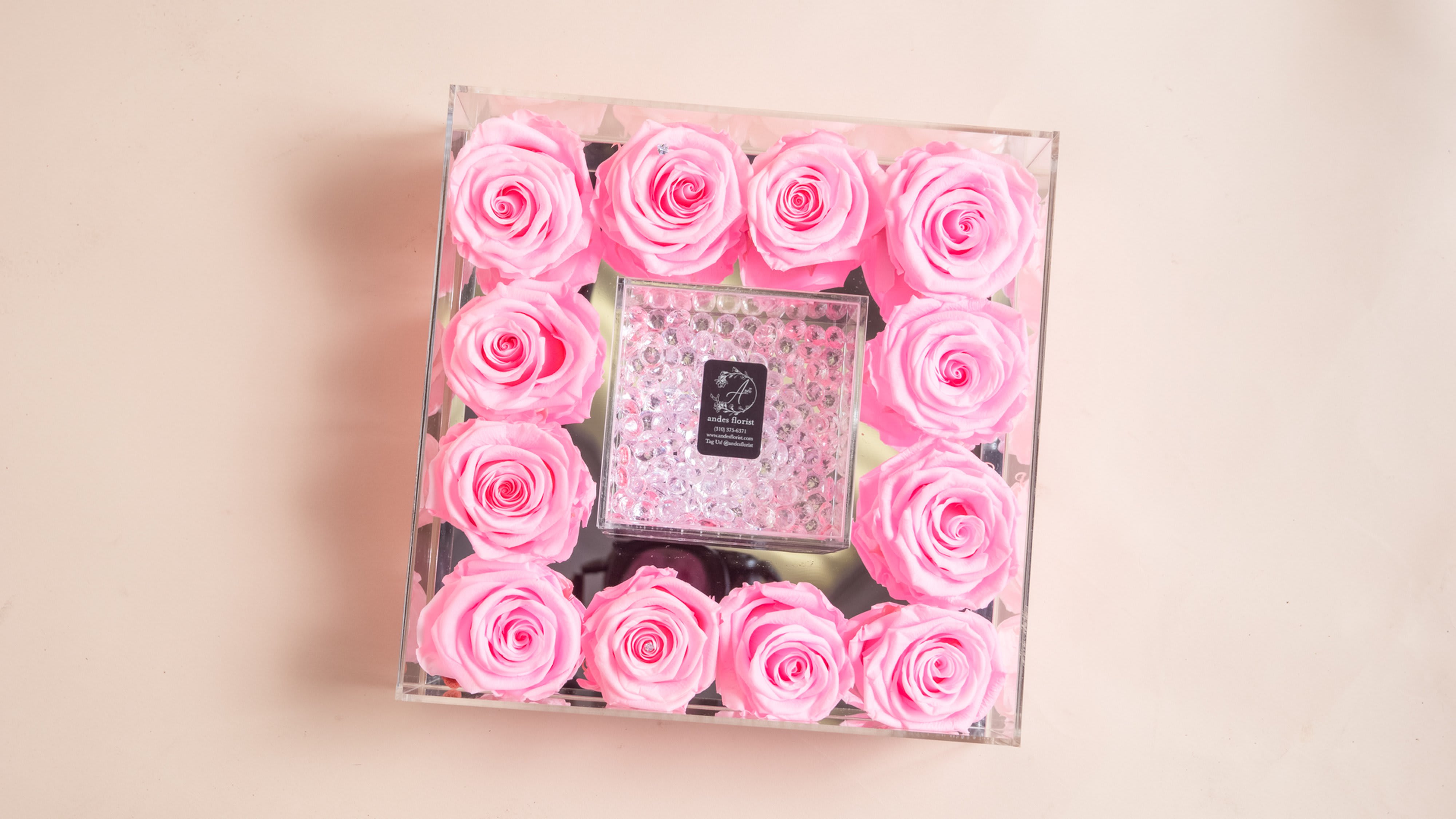 Pink roses arranged in a clear square box around a small center gift box.