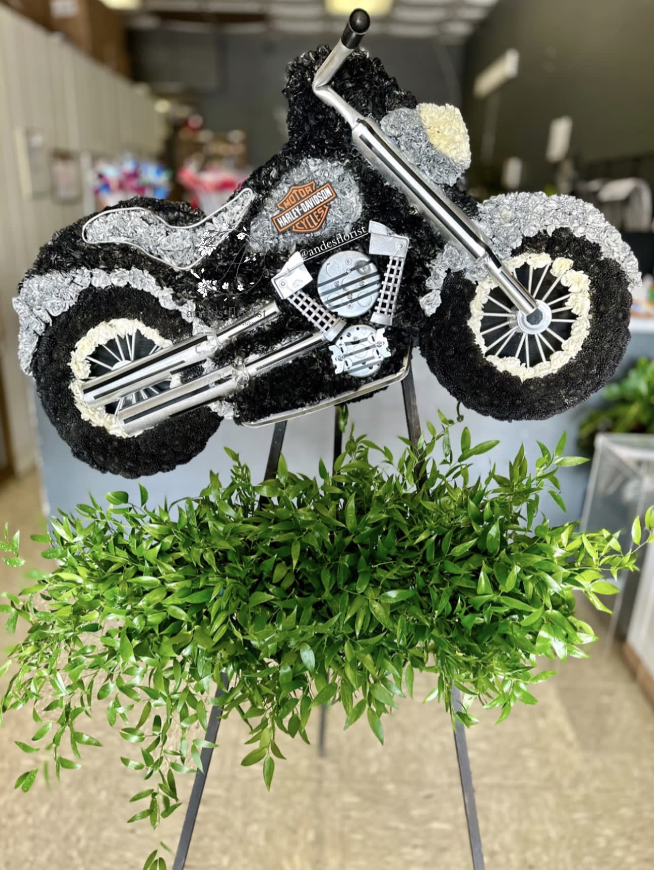 Motorcycle-shaped floral tribute with black, white, and gray flowers on an easel