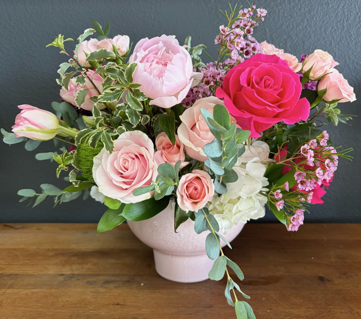 Blush Garden Serenade - A romantic and artfully gathered arrangement in soft blush, ivory, and vibrant pink tones, Blush Garden Serenade captures the beauty of a blooming spring garden. Featuring lush peonies, delicate garden roses, elegant spray roses, and charming wax flowers, this design is accented with fresh eucalyptus and textured greenery for a naturally graceful feel.  Designed in a modern ceramic vessel, this piece blends timeless elegance with a touch of whimsy—making it a heartfelt gift for celebrating the warmth, love, and beauty of motherhood. Perfect for brightening her day and filling her space with soft fragrance and joy.