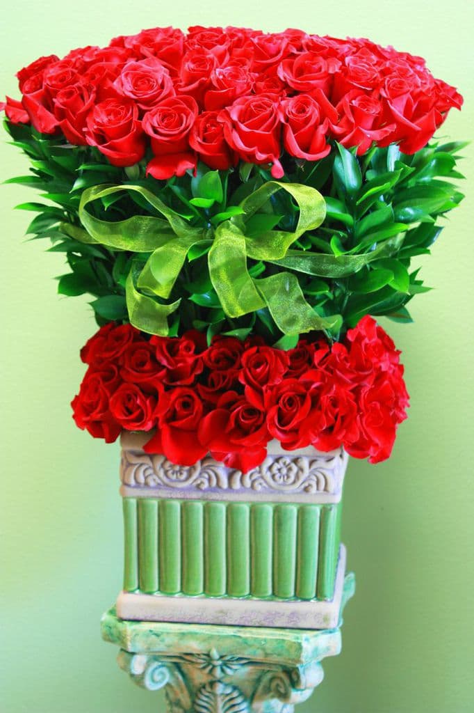 Tall arrangement of red roses with a green ribbon in a decorative pedestal vase