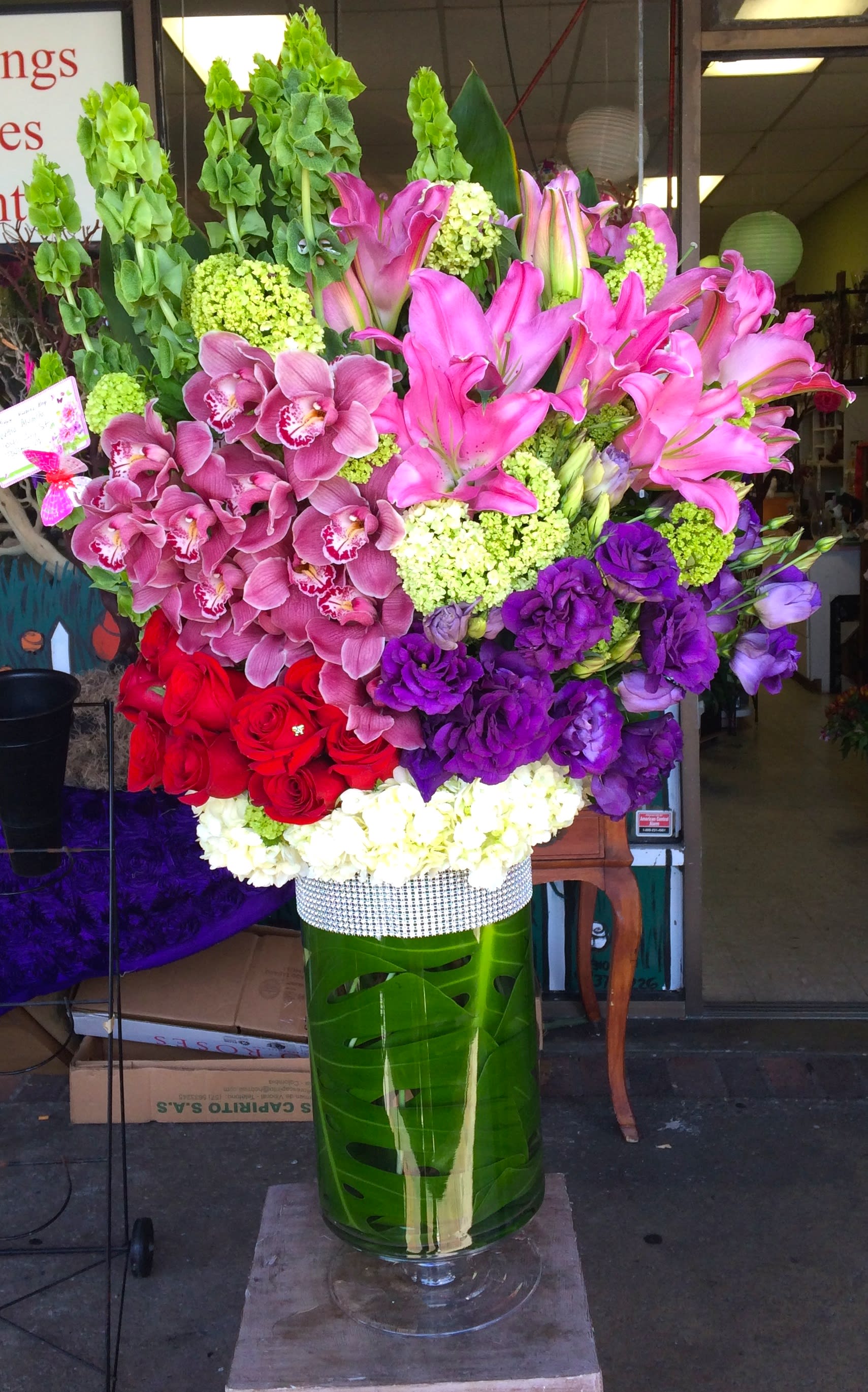 Tall mixed floral arrangement in a green vase with pink lilies, orchids, and red roses