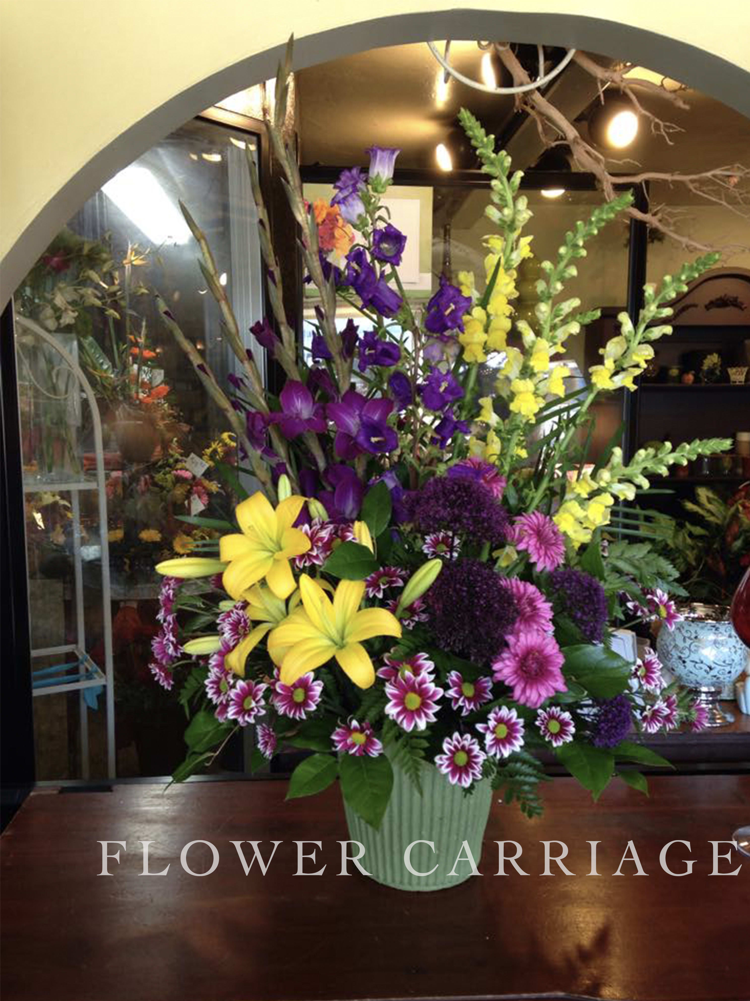 Tall mixed flower arrangement in a green vase with yellow lilies and purple blooms