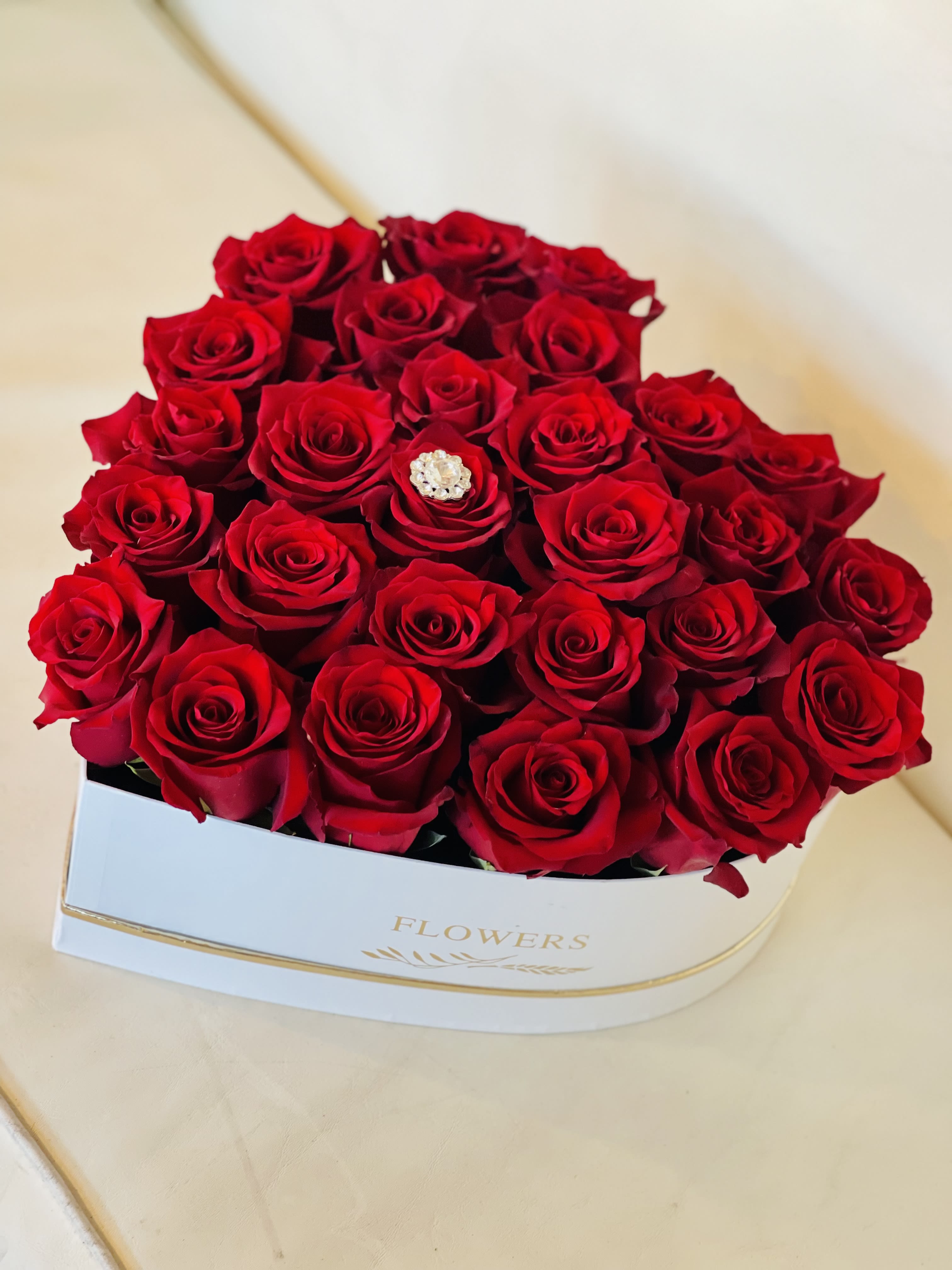   Baccarat Rouge  by Flower Carriage  - A sculpted heart of velvety red roses makes an unforgettable centerpiece for anniversaries, Valentine's Day, Mother's Day, or any "just because" moment when you want to say I love you. This flower arrangement heart is designed as a bold, romantic gift-tightly clustered red roses form a smooth, luxurious surface, and a single jeweled brooch at the center offers a sparkling focal point that catches the eye and elevates the whole piece.  Arranged in a crisp white box, the design pairs classic red and white contrast with rich texture; the roses read deep red in tone and sit low and compact for a polished, modern silhouette. Standard: H 5" x W 10" with approximately 12-18 roses; Deluxe: H 5" x W 12" with approximately 18-24 roses; Premium: H 6" x W 14" with approximately 24-30 roses. You can customize to any rose color-leave a note at checkout or call us if you prefer a different hue.  Perfect for the person you're thinking of-whether celebrating a birthday, graduation, new baby, wedding, or offering an apology, thank you, or get-well wish-this keepsake-worthy heart delivers an immediate feeling of being cherished. Place it on a bedside table or coffee table for an elegant, intimate display; order with confidence for seasonal occasions like Mother's Day or Breast Cancer Awareness Month and year-round moments of love and remembrance.