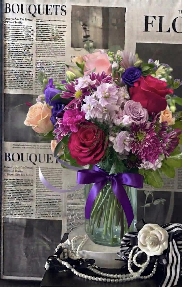 Mixed bouquet of pink, purple, and peach flowers in a glass vase with a purple ribbon
