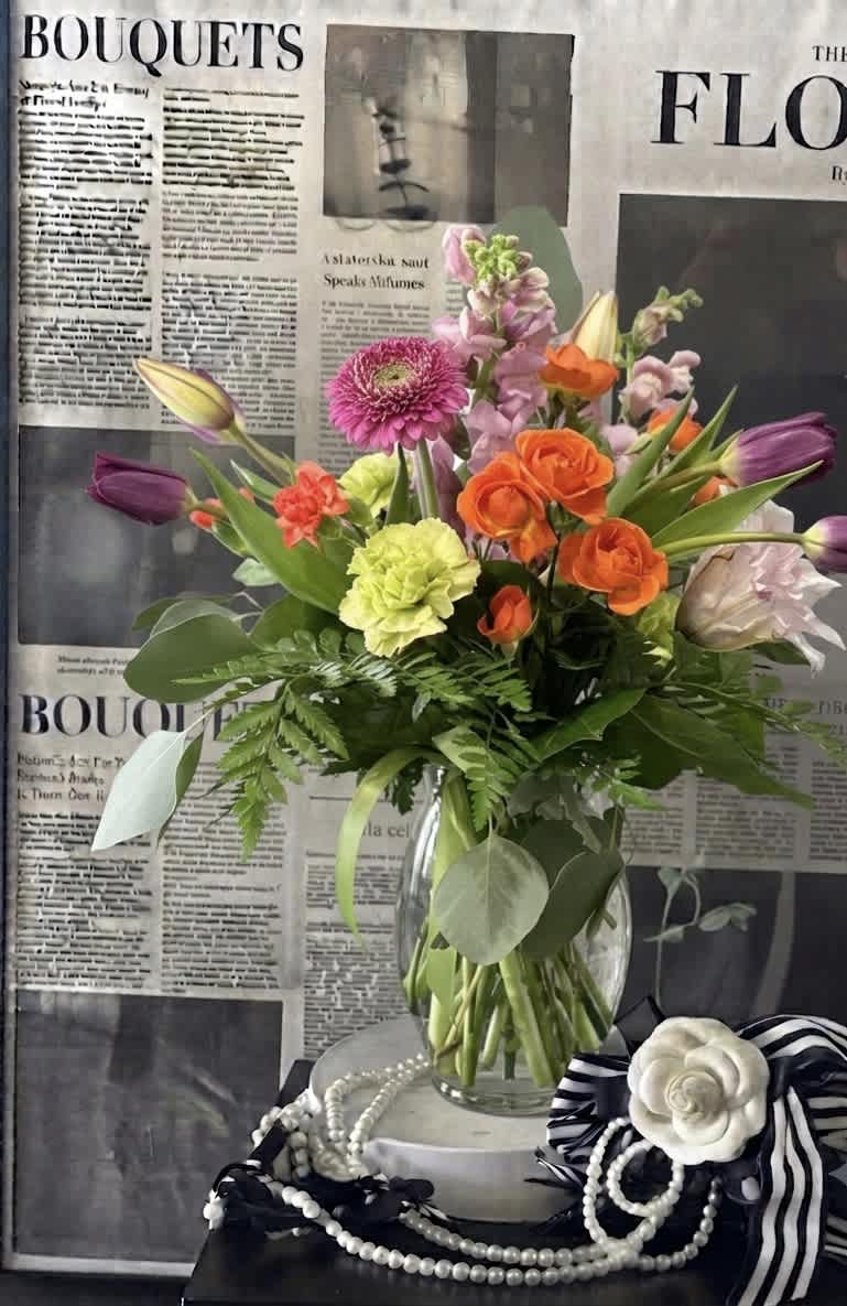 Mixed bouquet of orange, pink, purple, and yellow flowers in a clear glass vase