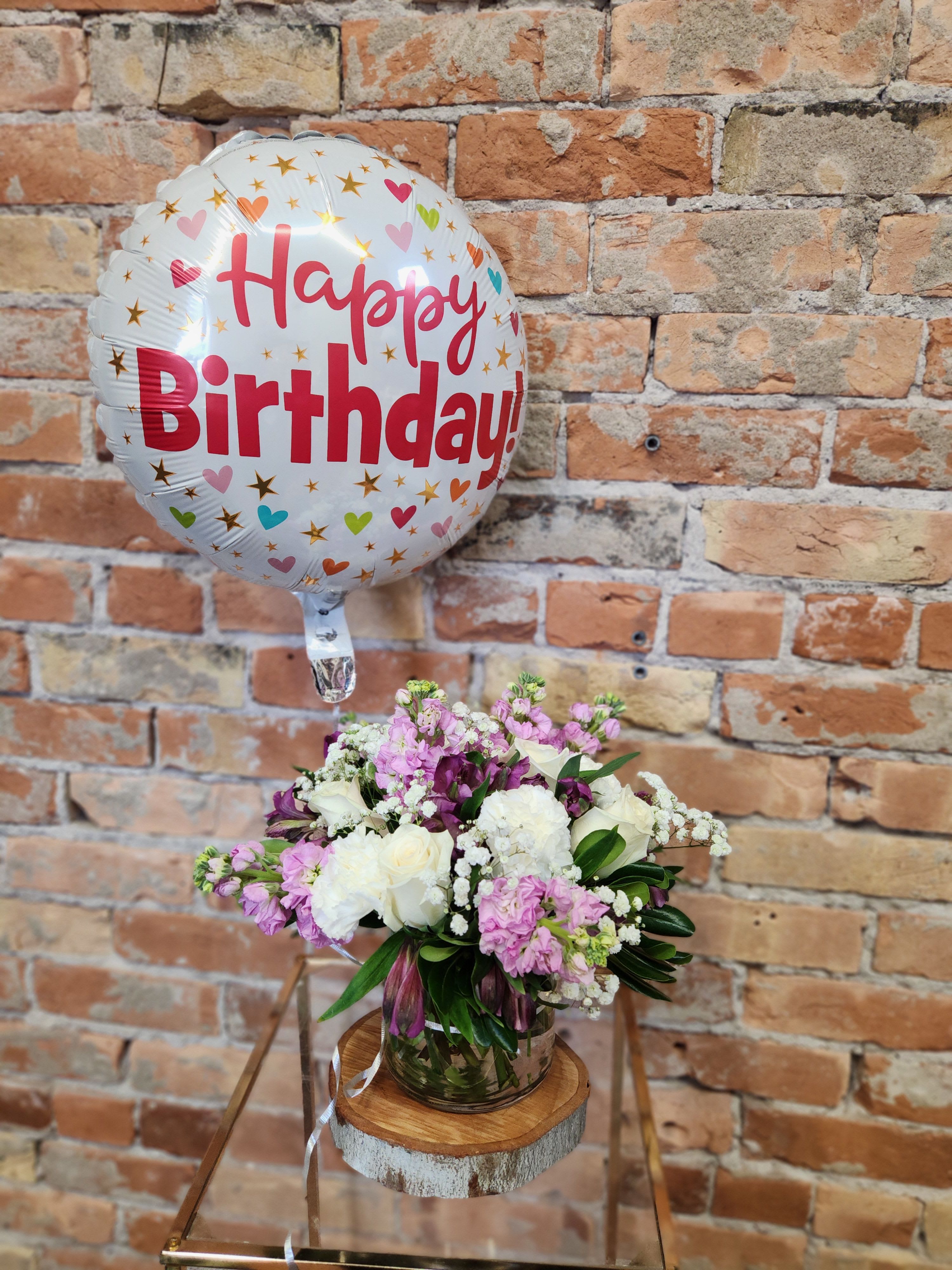 Birthday balloon and small pink and white flower arrangement in a clear vase on a stand.