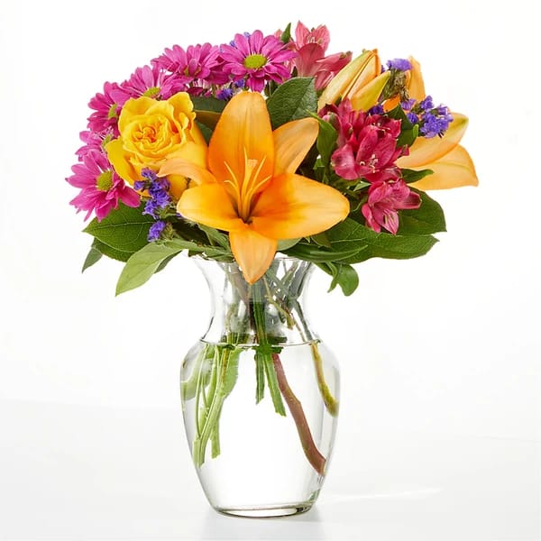 Bouquet of yellow lilies, pink daisies, and purple flowers in a clear glass vase