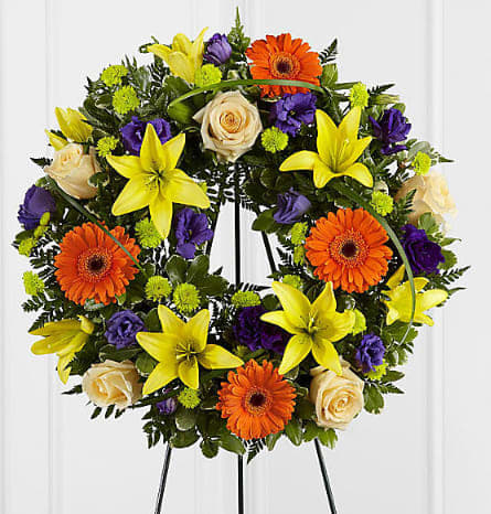Circular floral wreath with yellow lilies, orange gerberas, cream roses, and purple blooms