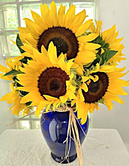 Summer Days - Big, sunlit sunflowers create an instant mood-lifter in this bright summer bouquet - a cheerful flower arrangement perfect for Father's Day, housewarming, or a just-because surprise. The rounded bouquet reads sunny and upbeat, the kind of gift that makes the person you're thinking of feel noticed and genuinely cheered.  Several large sunflowers with vivid yellow petals and dark centers are gathered tightly and presented in a glossy cobalt-blue glass vase, the bold yellow-and-blue contrast making a striking casual centerpiece. A simple raffia ribbon ties the look together for a rustic, homey touch; the compact, rounded shape works well on tabletops, kitchen counters, or an office desk.  Ideal for summer celebrations yet equally suited to spring, autumn, Thanksgiving, get-well wishes, or an "I'm sorry" gesture, this arrangement sends a warm, optimistic message across seasons. Vase included; no special care needed beyond fresh water and a cool spot away from direct midday sun to keep those big faces looking bright.