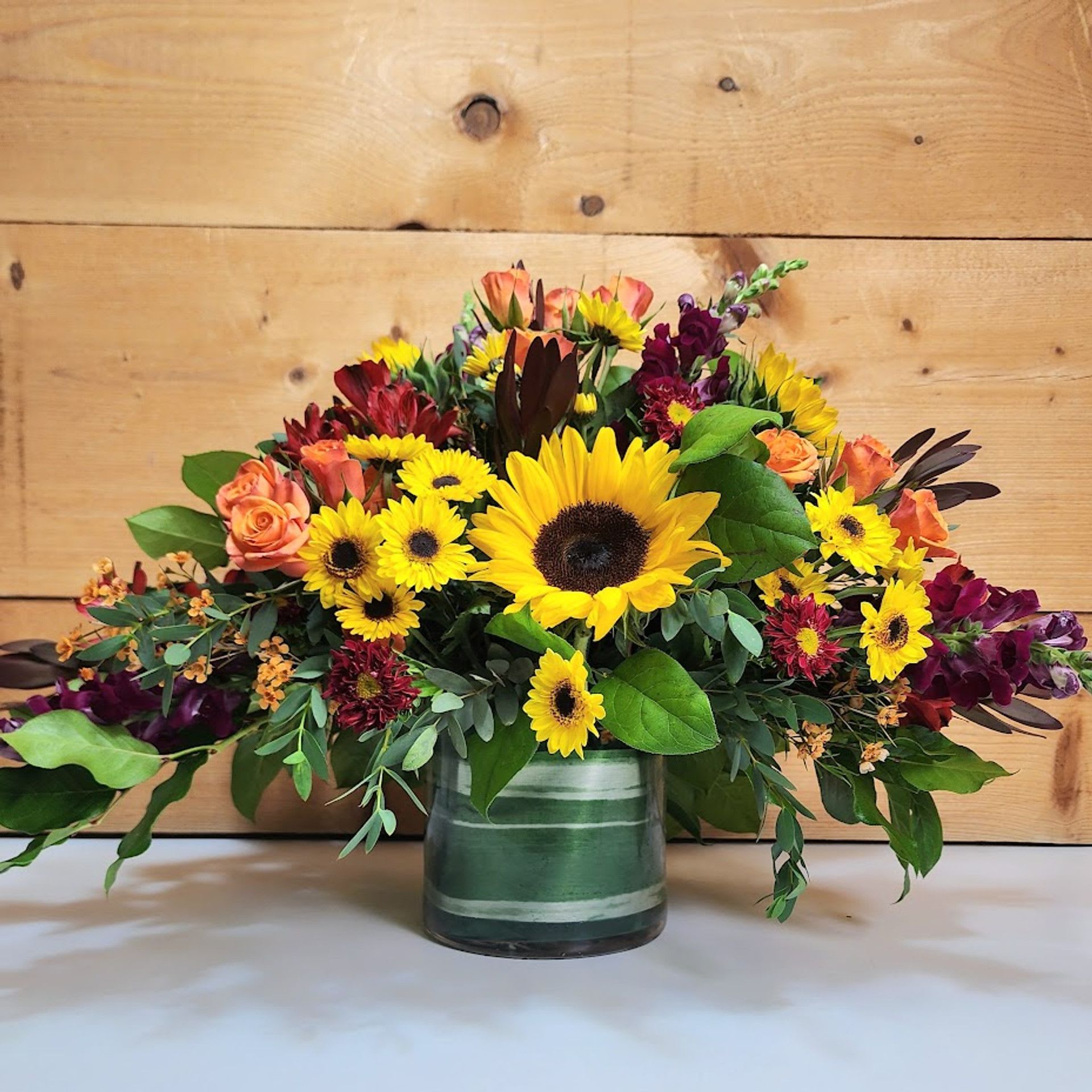 Autumn Sunshine Centerpiece - Autumn Sunshine Centerpiece  Bright and bold, this centerpiece captures the essence of fall’s beauty. Featuring radiant sunflowers and golden yellow daisies alongside orange spray roses, deep burgundy mums, and red alstroemeria, it’s accented with safari sunset, snapdragons, and lush greenery for texture and fullness. Designed in a low, wide container, this vibrant display is perfect for a dining table, buffet, or gathering space — bringing warmth, cheer, and seasonal charm to any celebration.