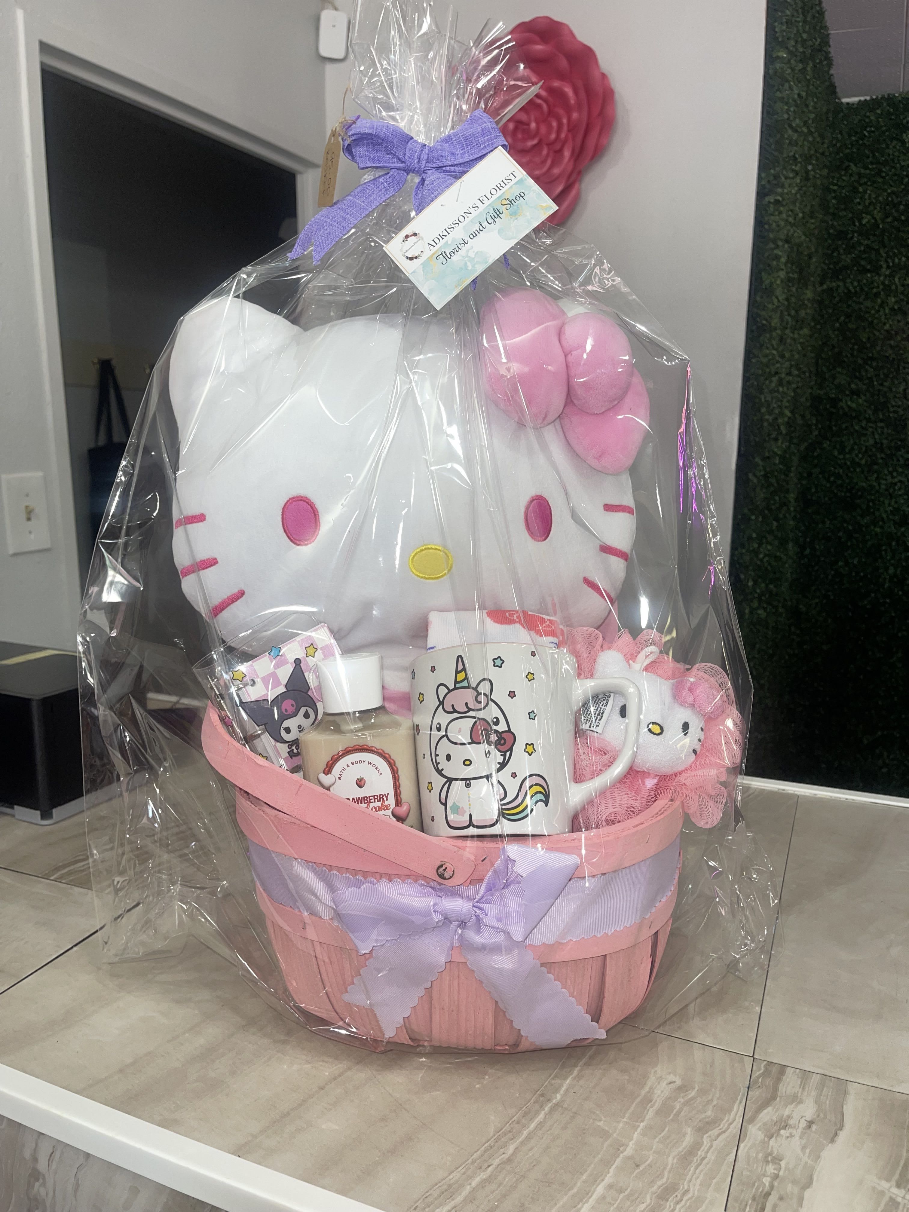 Hello Kitty Basket - A cheerful Hello Kitty gift basket that feels like a hug in pink, white, and lavender - perfect as a birthday bouquet alternative, a get-well surprise, or a just-because delight for any Hello Kitty fan. This character-themed flower arrangement substitute is designed to make the recipient feel playfully celebrated: the plush Hello Kitty perched on top gives immediate charm, while the soft color palette reads sweet and youthful for children or nostalgic adults.  The basket is presented in a pink woven container, wrapped in clear cellophane and finished with a lavender ribbon. Inside you'll find a large white (and/or pink) Hello Kitty plush, a matching character mug, a body lotion bottle, a scented candle, packaged treats, a small decorative item, and a gift card - all arranged around light floral accents in pink, white, and purple tones to keep the look fresh and feminine. Plastic and cellophane wraps secure smaller items so everything arrives neat and gift-ready.  Choose this for birthdays, get-well wishes, or a spontaneous token of affection when you want her to feel seen and smiled at; it's the kind of gift that brightens a room and lingers as a keepsake. Available in one presentation size as shown; care is limited to removing any cellophane before displaying the plush and candle, and following label instructions on the body lotion and candle.