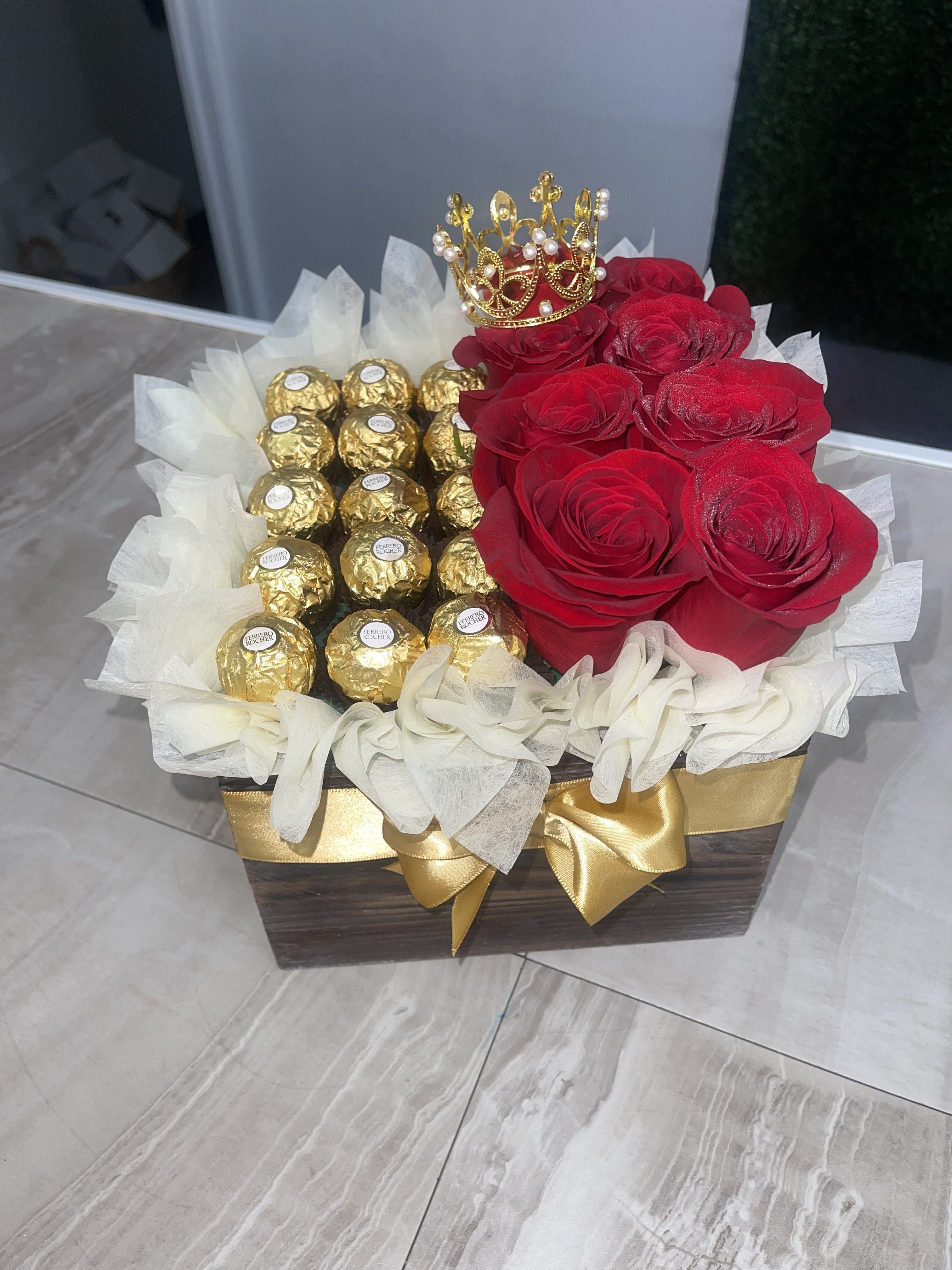 Roses Box Combo - A cluster of deep red roses paired with gold-wrapped chocolates makes a bold, celebratory statement - perfect as a romantic birthday gift.The mix of red, gold satin ribbon reads festive and thoughtful, so the recipient will feel seen and celebrated.  This arrangement pairs red roses with rows of Ferrero Rocher and other gold-wrapped chocolate candies tucked into a dark wood-look box, cushioned by white decorative tissue paper. Finish details include a gold satin ribbon decorative tissue. The box is compact and gift-ready, combining floral fragrance with the indulgence of premium chocolates.  Ideal for  a special birthday, or any occasion where you want to say "thanks" with both flowers and sweets, this keepsake-style wooden box is designed for immediate display and enjoyment. No special care beyond fresh water and a cool spot is required for the roses; order early to ensure availability and to have this playful, refined gift arrive fresh for the moment that matters.