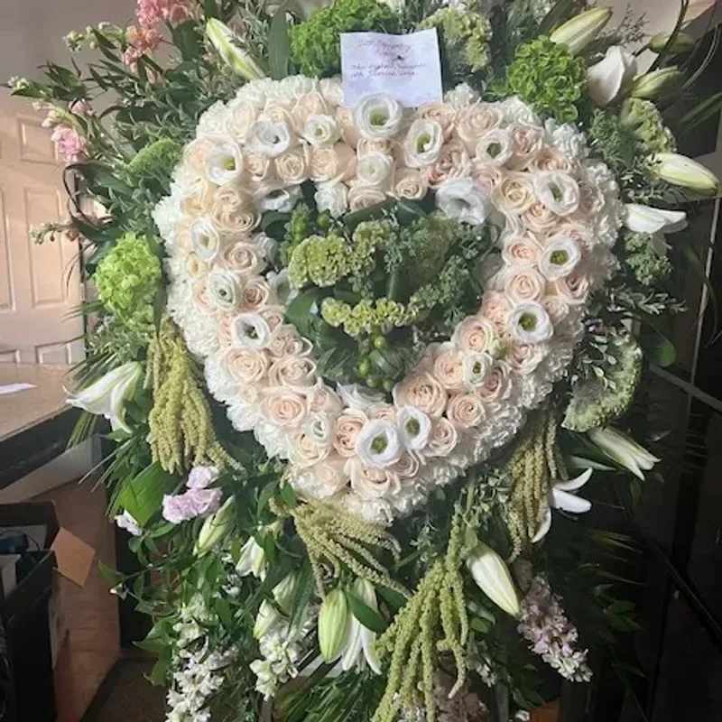 fhfh - A large heart-shaped sympathy wreath crafted from tightly packed cream and blush roses creates an immediate impression of gentle reverence-the perfect floral arrangement for a memorial tribute or a serene wedding display. The white lilies woven into the outer edge and the pale green accents give a soft, calming palette that reads both romantic and solemn, helping the person you're honoring feel remembered and seen.  Built on a floral wreath frame, the center is filled with layered greenery and small clustered blooms for a dense, formal look that holds its shape during services or display. Colors are white, pink and green; flowers include roses, lilies and complementary seasonal blooms, arranged for a full, textured appearance that reads beautifully from a distance and up close. This heart-shaped design is sized for a prominent visual presence and arrives ready to place on an easel or hang, with typical care being kept cool and out of direct sun to preserve freshness.