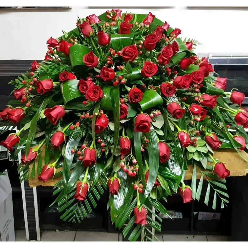 Casket Spray 1 - A lush, formal spray of deep red roses set against broad glossy green foliage creates a dignified sympathy and funeral tribute that reads as both respectful and richly emotional. This casket spray is arranged in a full, cascading shape so the person you're honoring will be remembered with a dramatic, polished presentation-an appropriate choice for memorial services, anniversaries of loss, or when you want to send a solemn, heartfelt message.  The arrangement features a dense mass of red roses layered with tropical, palm-like leaves and smaller textured greenery for contrast; small red berry accents are tucked throughout to add depth and refined detail. Its abundant, wide-spreading silhouette offers strong visual impact from the front and sides, and the combination of deep crimson and glossy green delivers a timeless, formal look that complements traditional funeral settings.